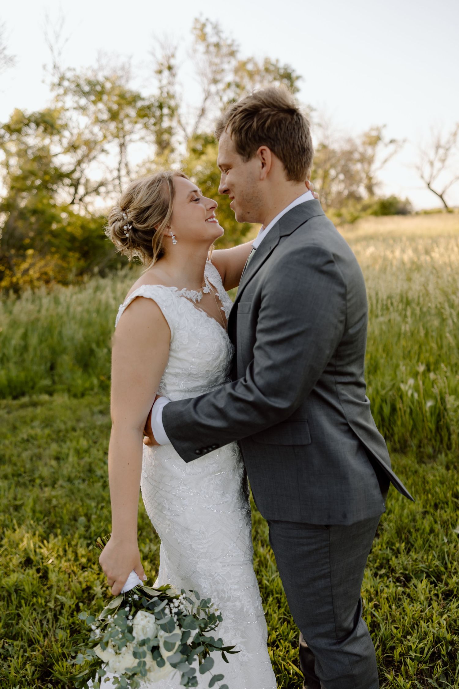 A romantic wedding couple embraces in a sunlit meadow with tall grass and trees in the background.