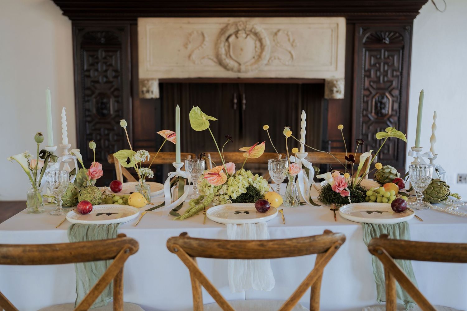 Elegant wedding reception tablescape with floral centerpieces, candles, and fruit display in front of ornate fireplace.