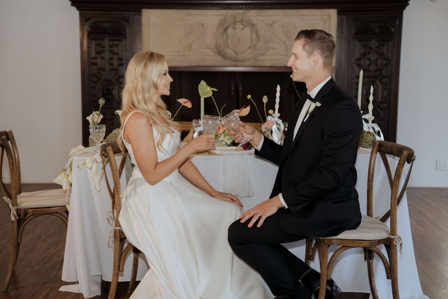 Elegant bride and groom in wedding attire sitting by fireplace, sharing a toast at decorated reception table.