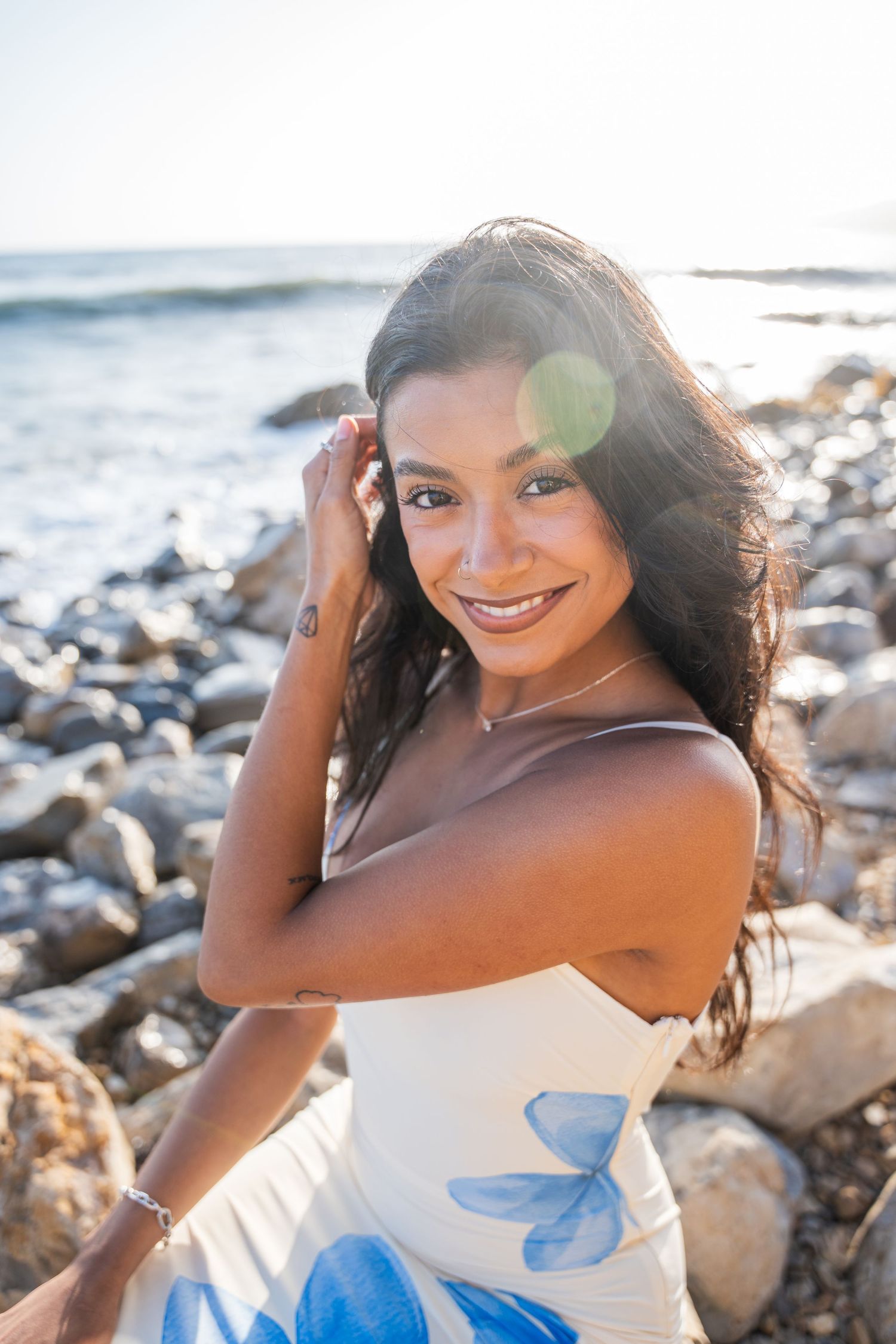 Smiling woman in white floral dress sitting on rocky beach with ocean and sunlight in background.
