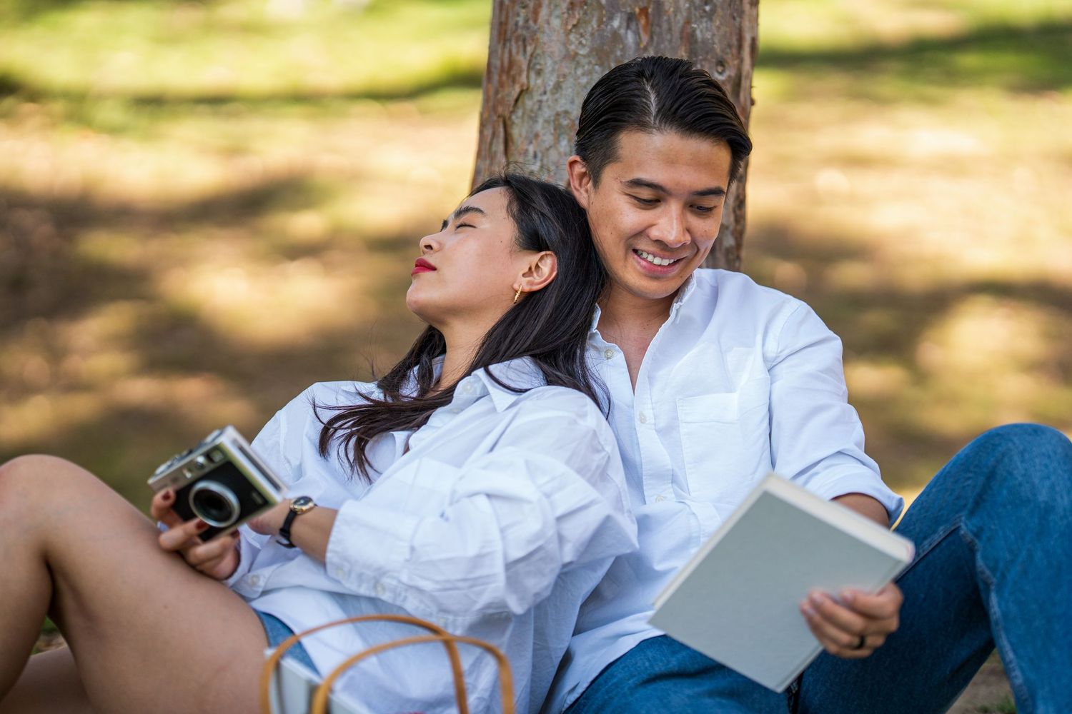 Young Asian couple relaxing under tree in park, woman with camera leaning on man reading a book.