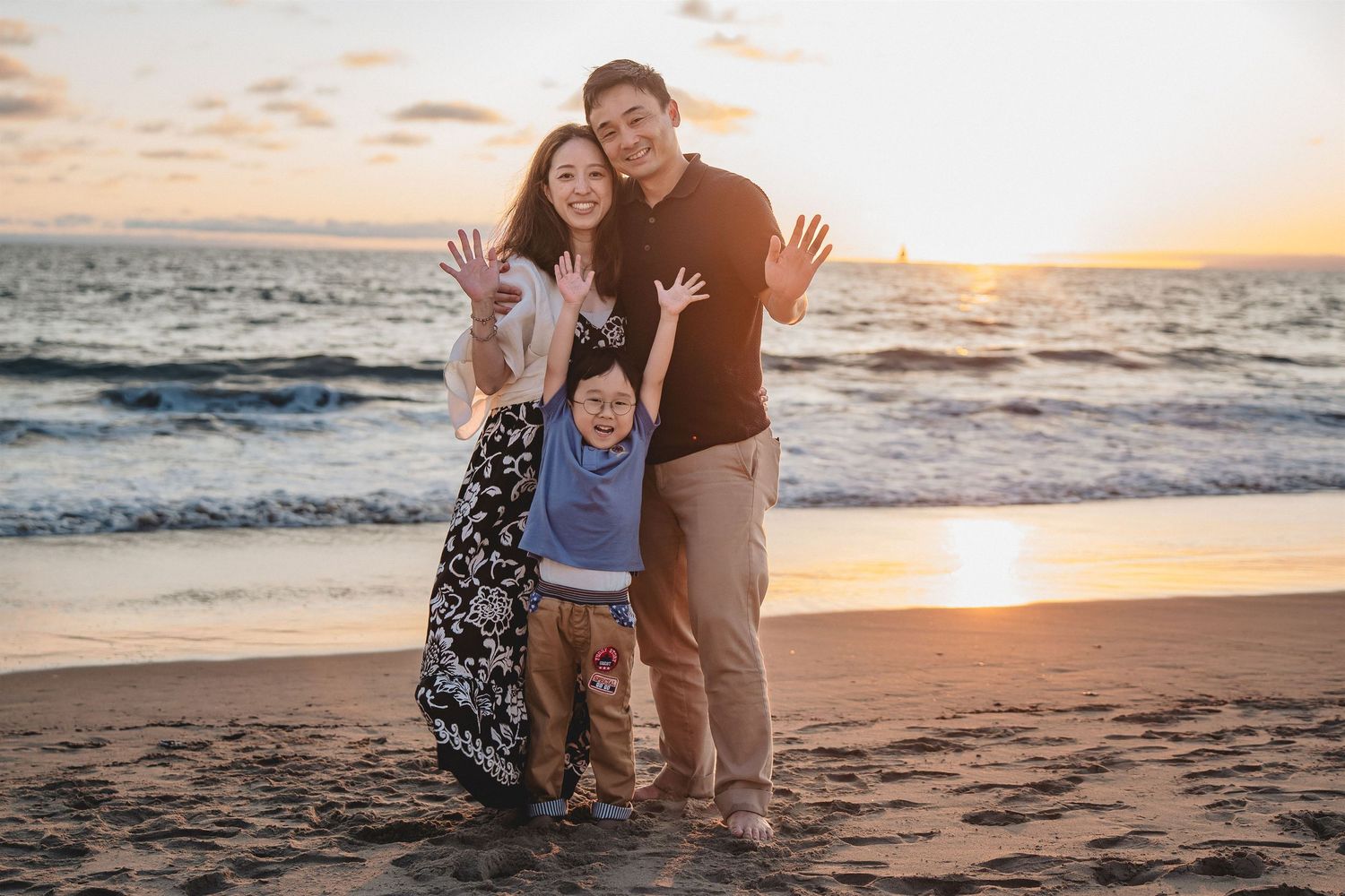 Family of three waving and smiling on a beach at sunset, with ocean waves in the background. Hermosa beach pier