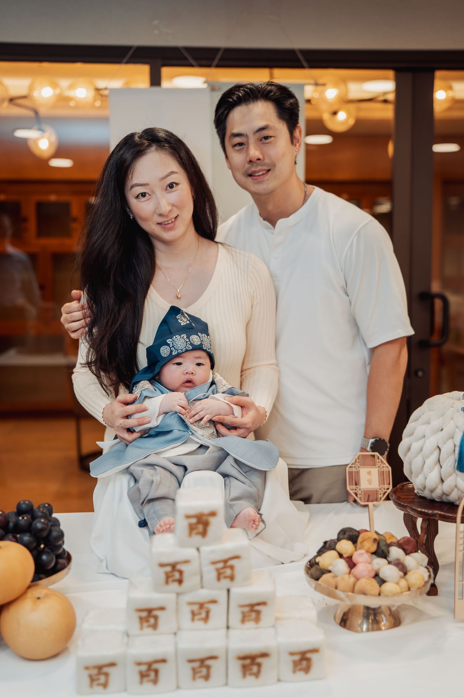 Asian family celebrating baby's 100-day milestone with traditional Korean doljabi ceremony setup featuring fruit and rice cakes.