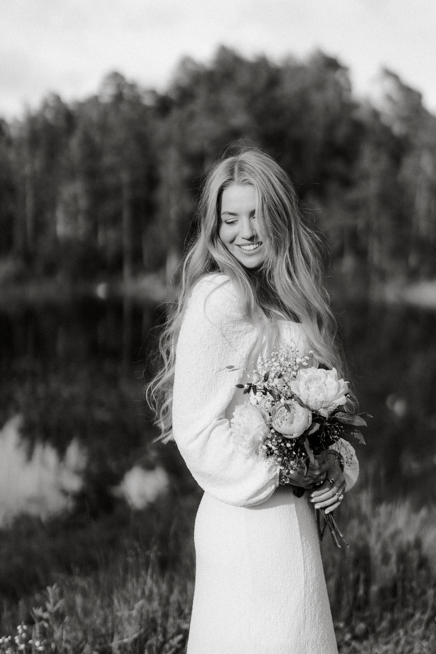 Black and white portrait of smiling blonde bride holding bouquet near reflective lake.