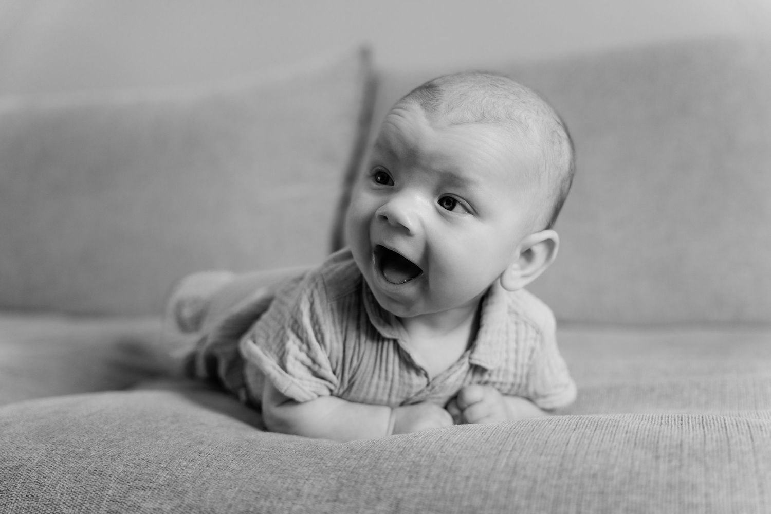 Black and white photo of a happy baby lying on tummy on a couch, looking up with mouth open.