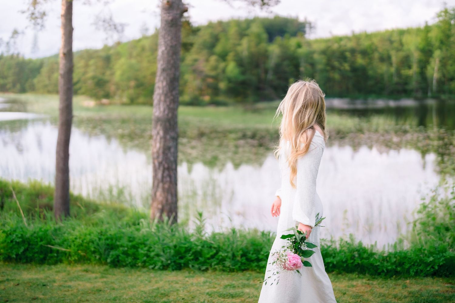 Bride in white gown holding pink flowers by a serene lake surrounded by lush green trees.