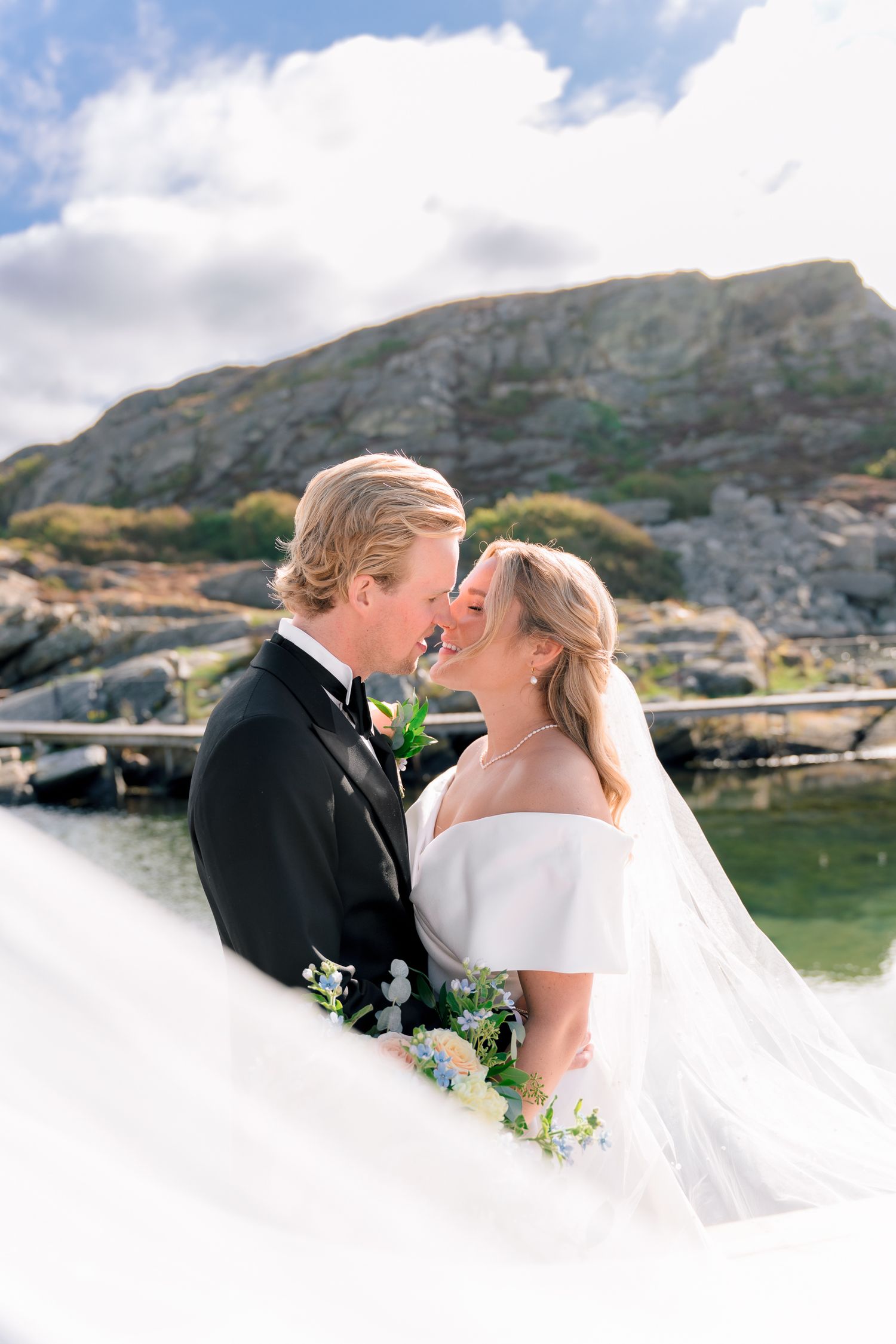 Bride and groom touch foreheads romantically as her veil flows in the breeze by a rocky shore.