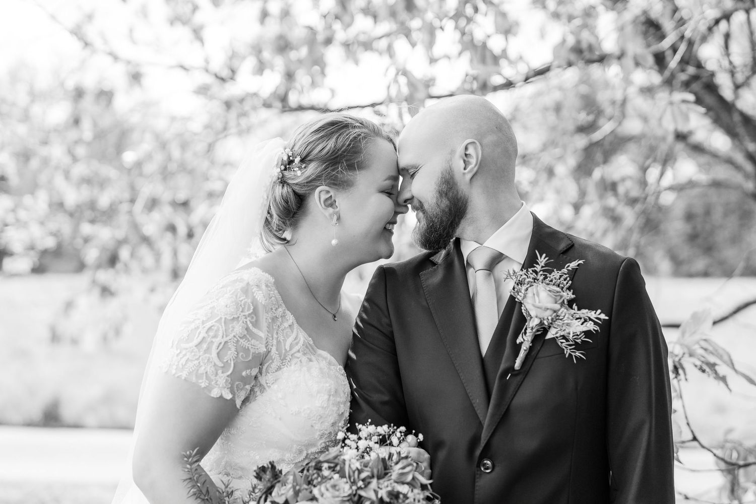 Black and white wedding photo of bride and groom touching foreheads tenderly outdoors, groom in suit with boutonniere.