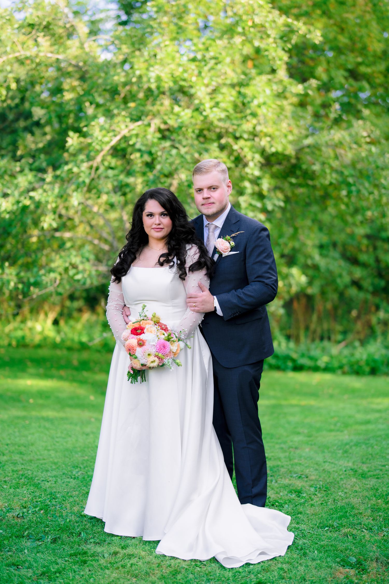 Bride in white gown holding pink bouquet stands with groom in dark suit on green lawn with trees.