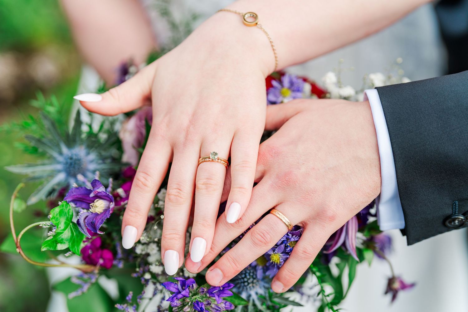 Bride and groom showing wedding rings on bouquet of purple flowers.
