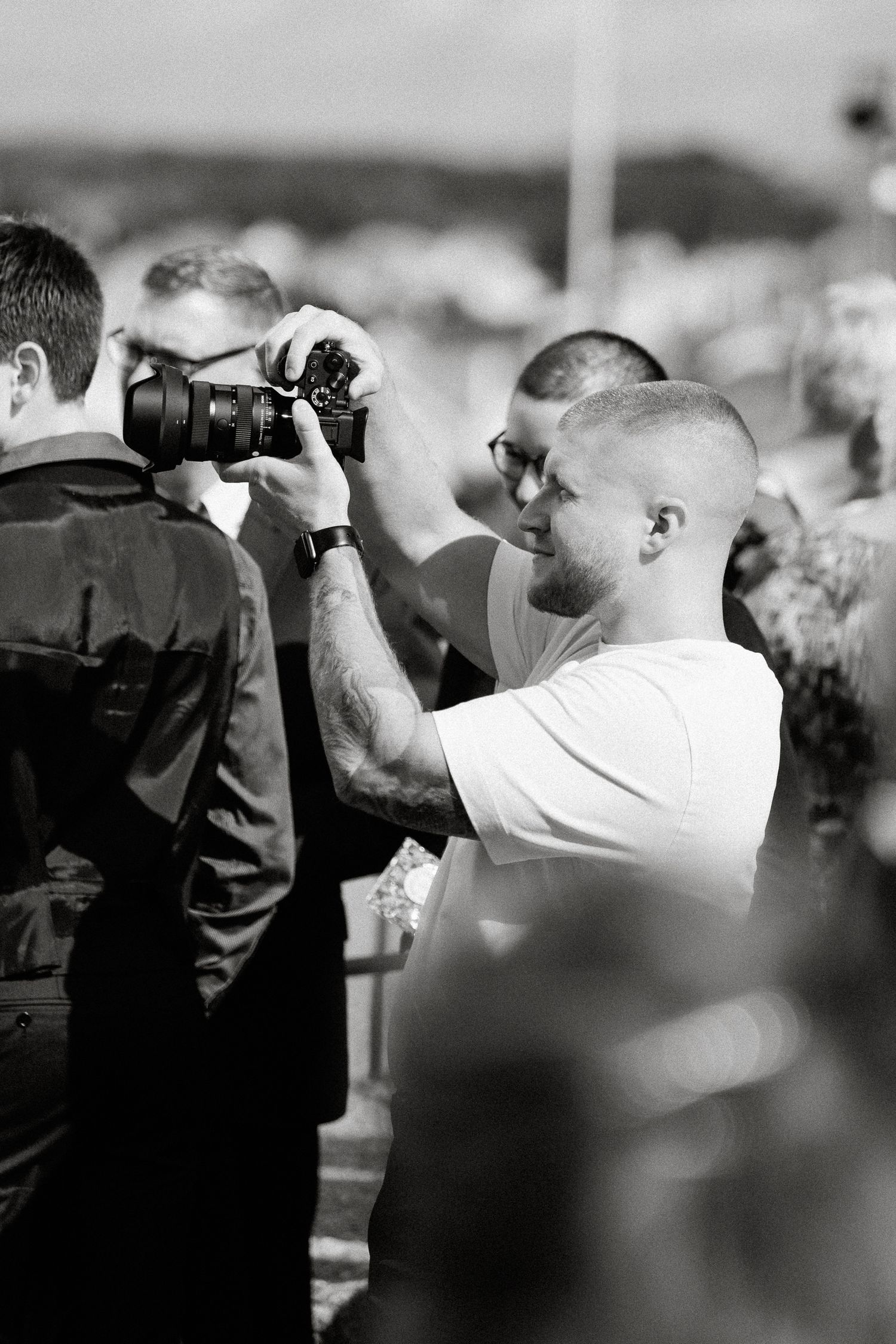 Black and white photo of a tattooed man holding a camera at an outdoor event.