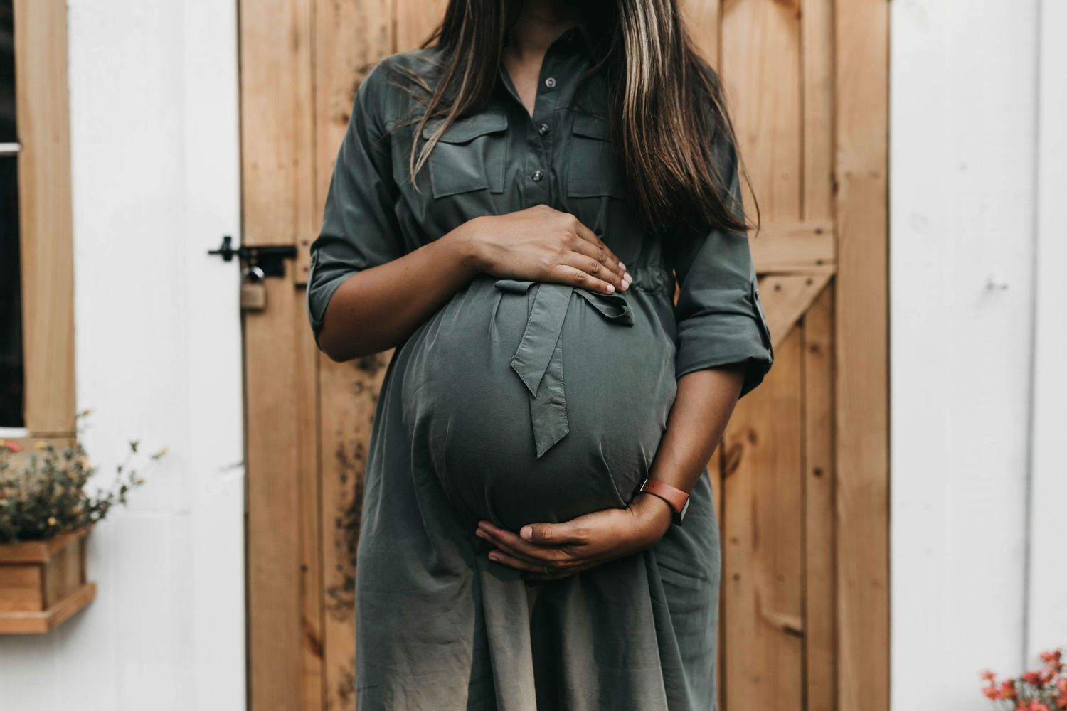 Pregnant woman in olive green dress cradling her baby bump outdoors near a rustic wooden door.