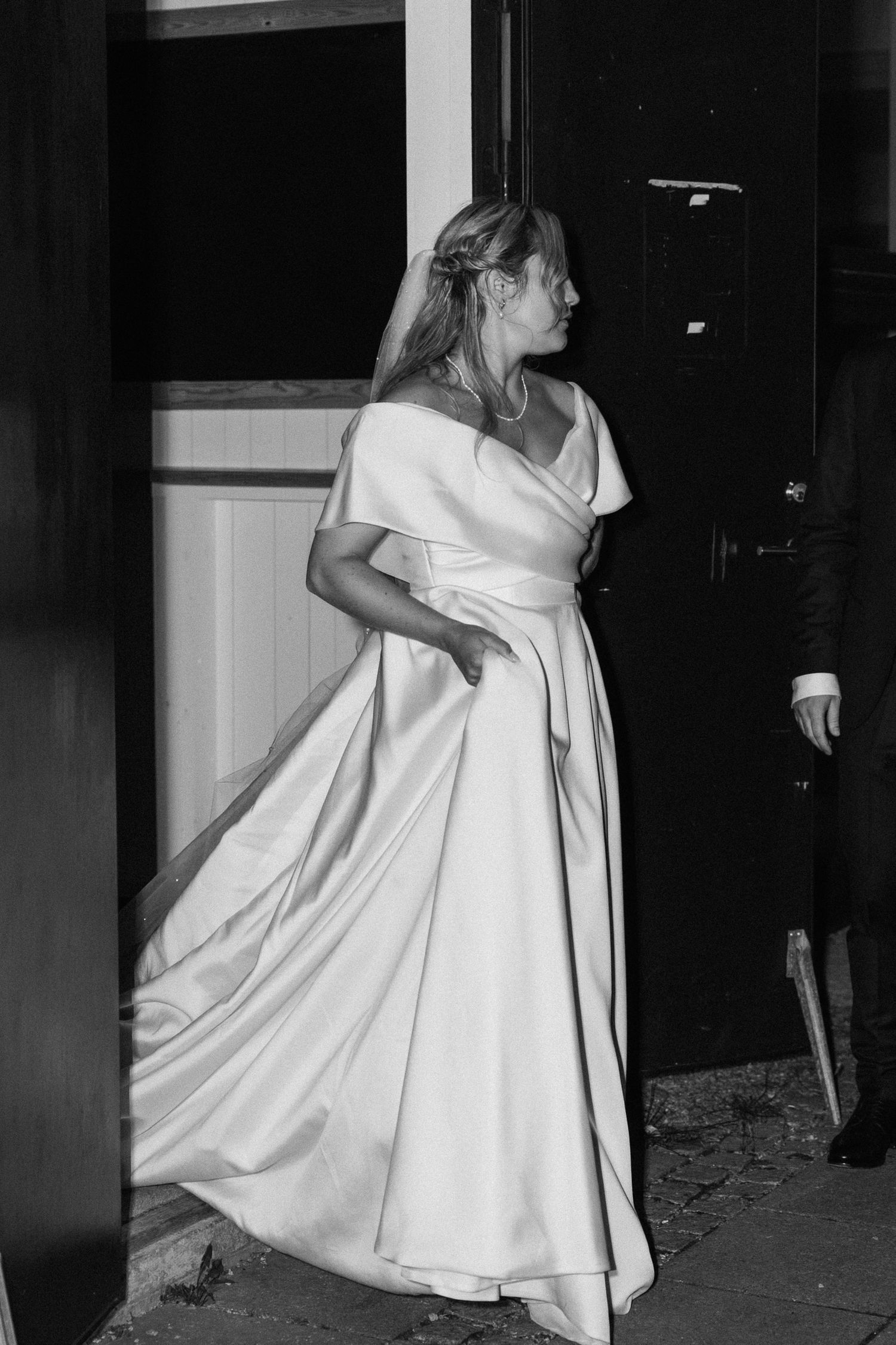 Black and white portrait of elegant bride in off-shoulder satin gown standing in doorway.