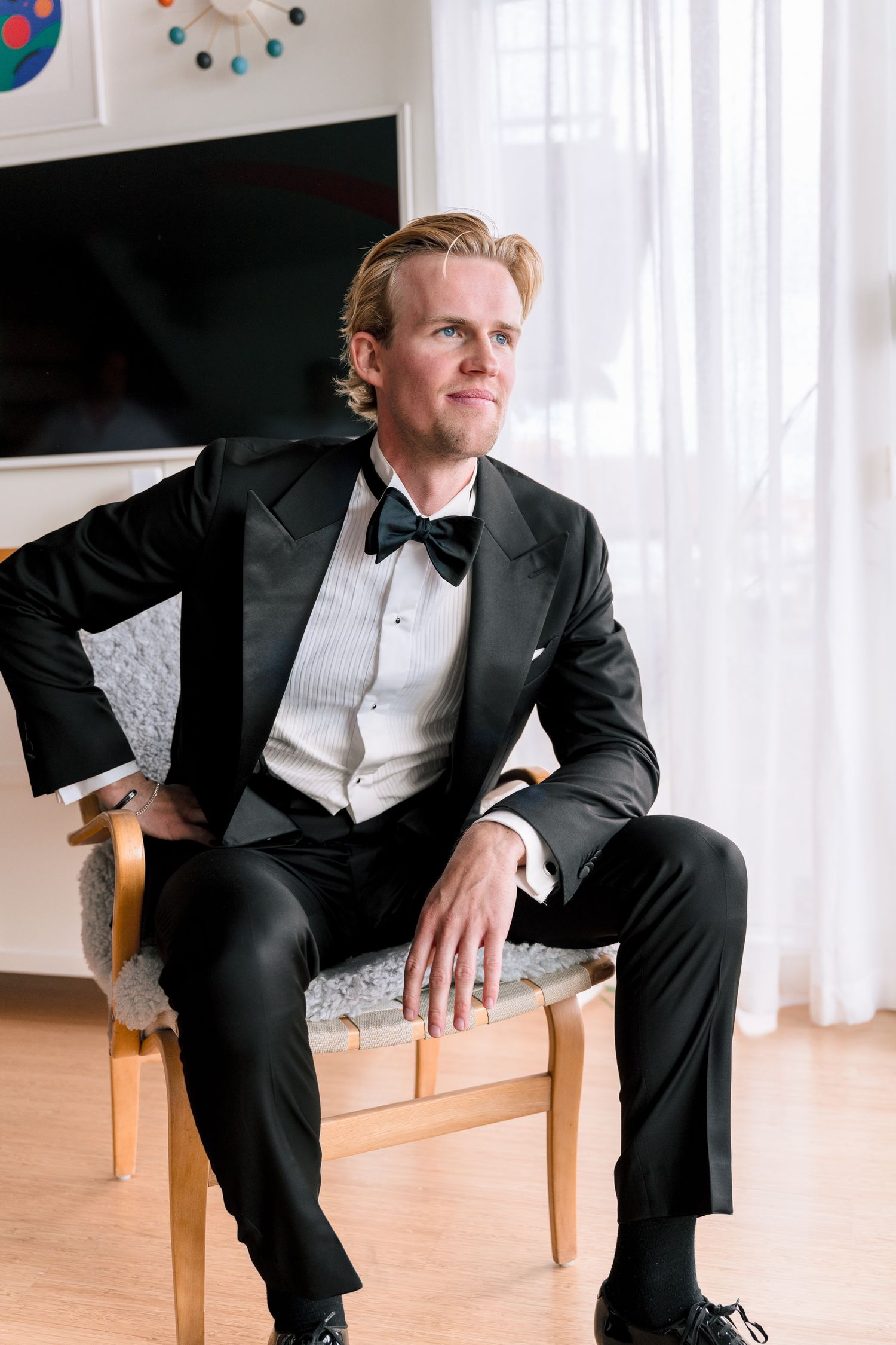 Handsome groom in classic black tuxedo with bow tie sitting in chair by window on wedding day.
