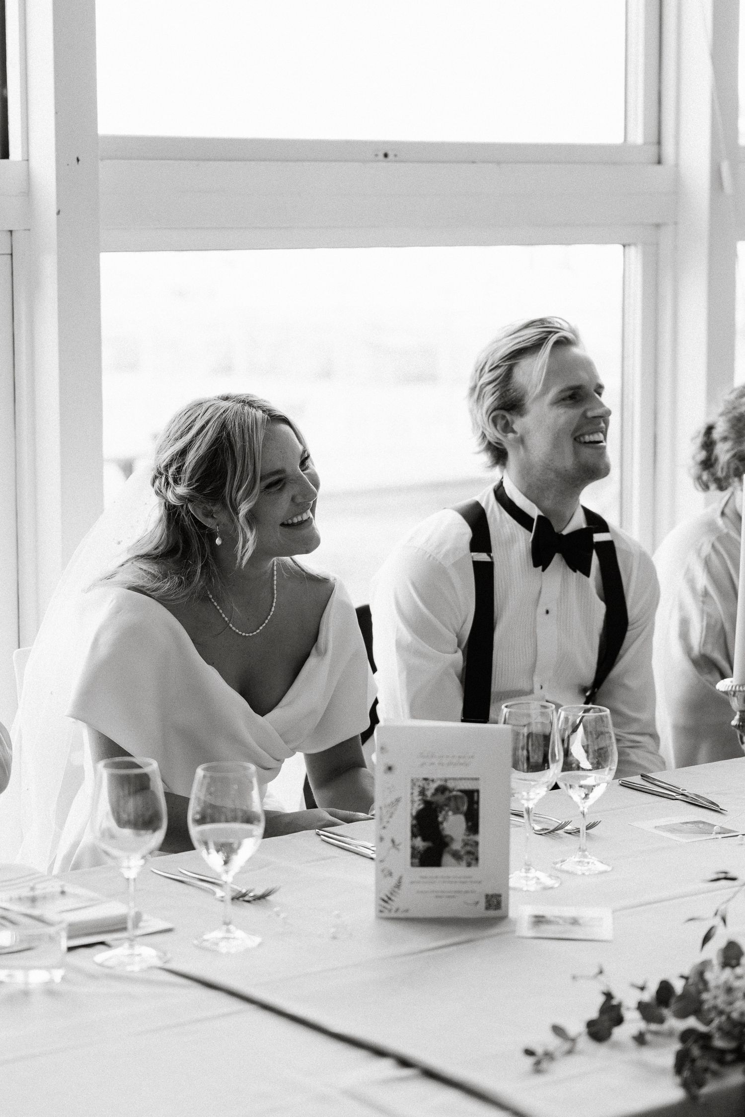 Black and white photo of a smiling bride and groom sitting at their wedding reception table with glasses and decor.