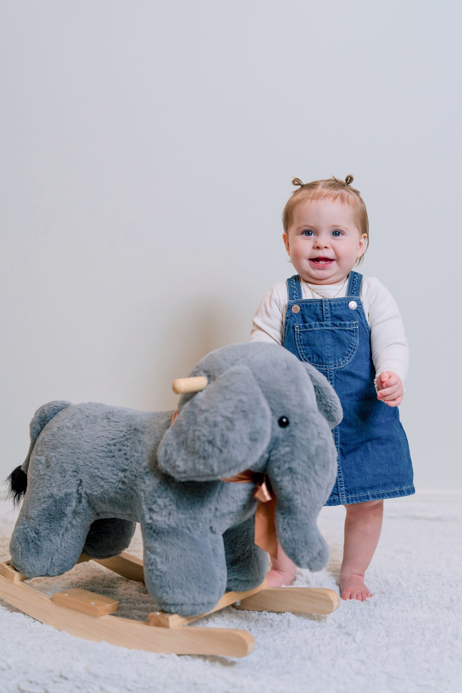 Smiling baby girl in denim overalls standing next to a gray plush elephant rocking toy on a white carpet.