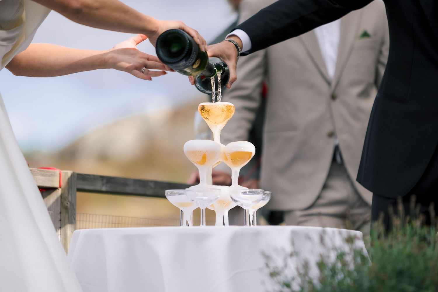Bride and groom pouring champagne into a elegant tower of glasses at their wedding reception outdoors.
