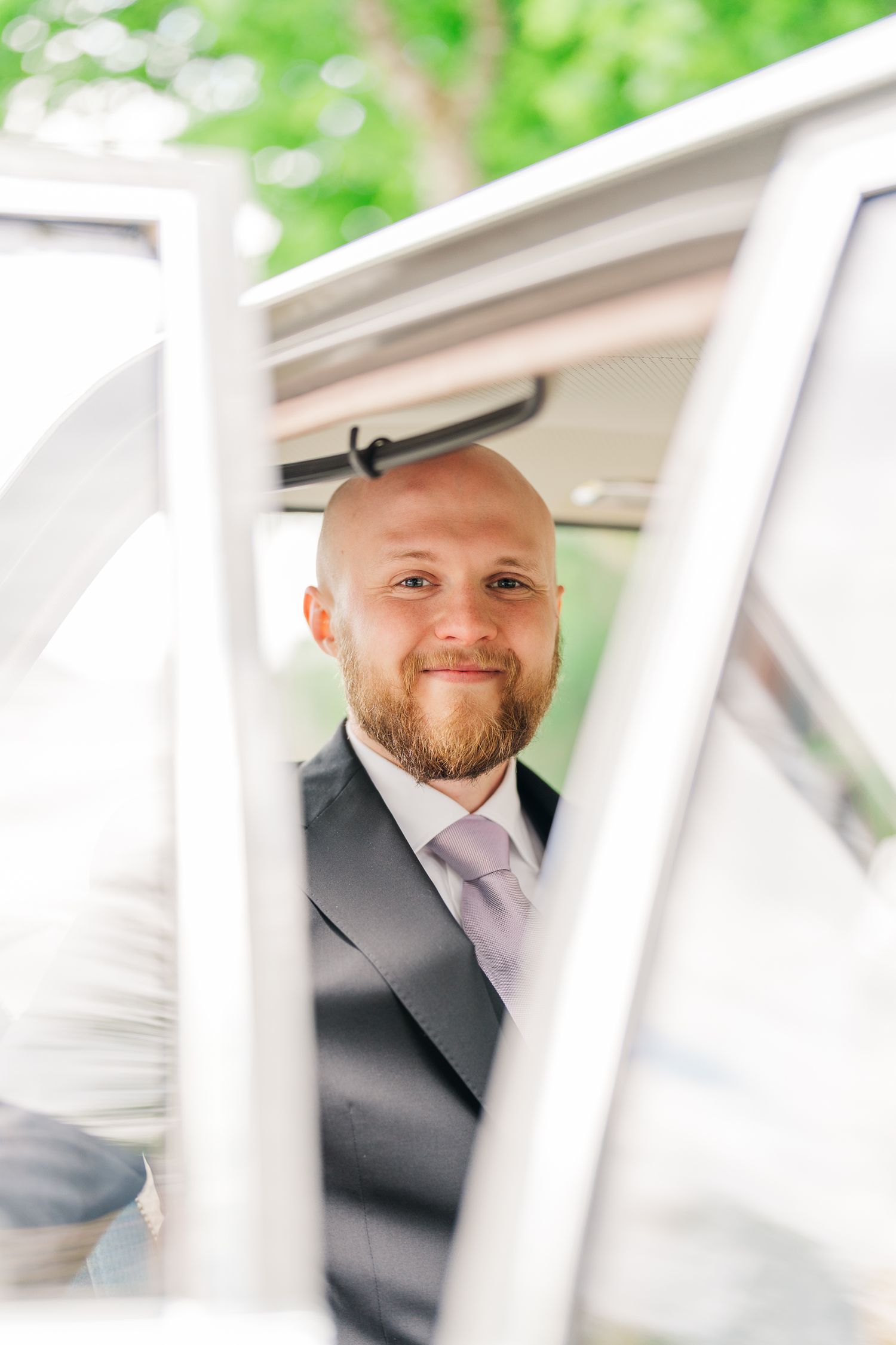 Smiling bald groom with beard wearing dark suit and lavender tie, photographed through a white car window.