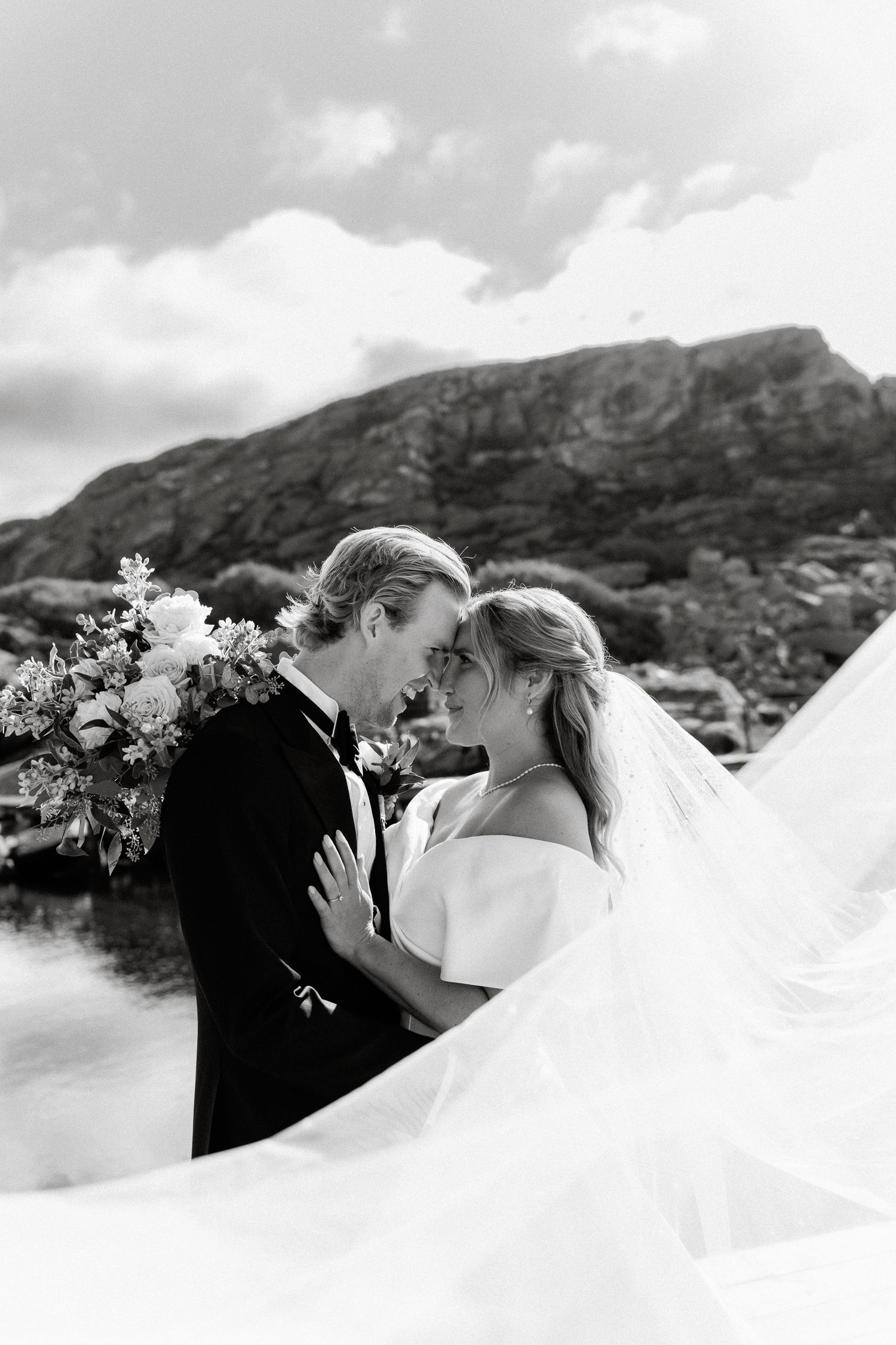 Black and white portrait of bride and groom nose-to-nose, dramatic veil sweep, rocky mountain lake background.