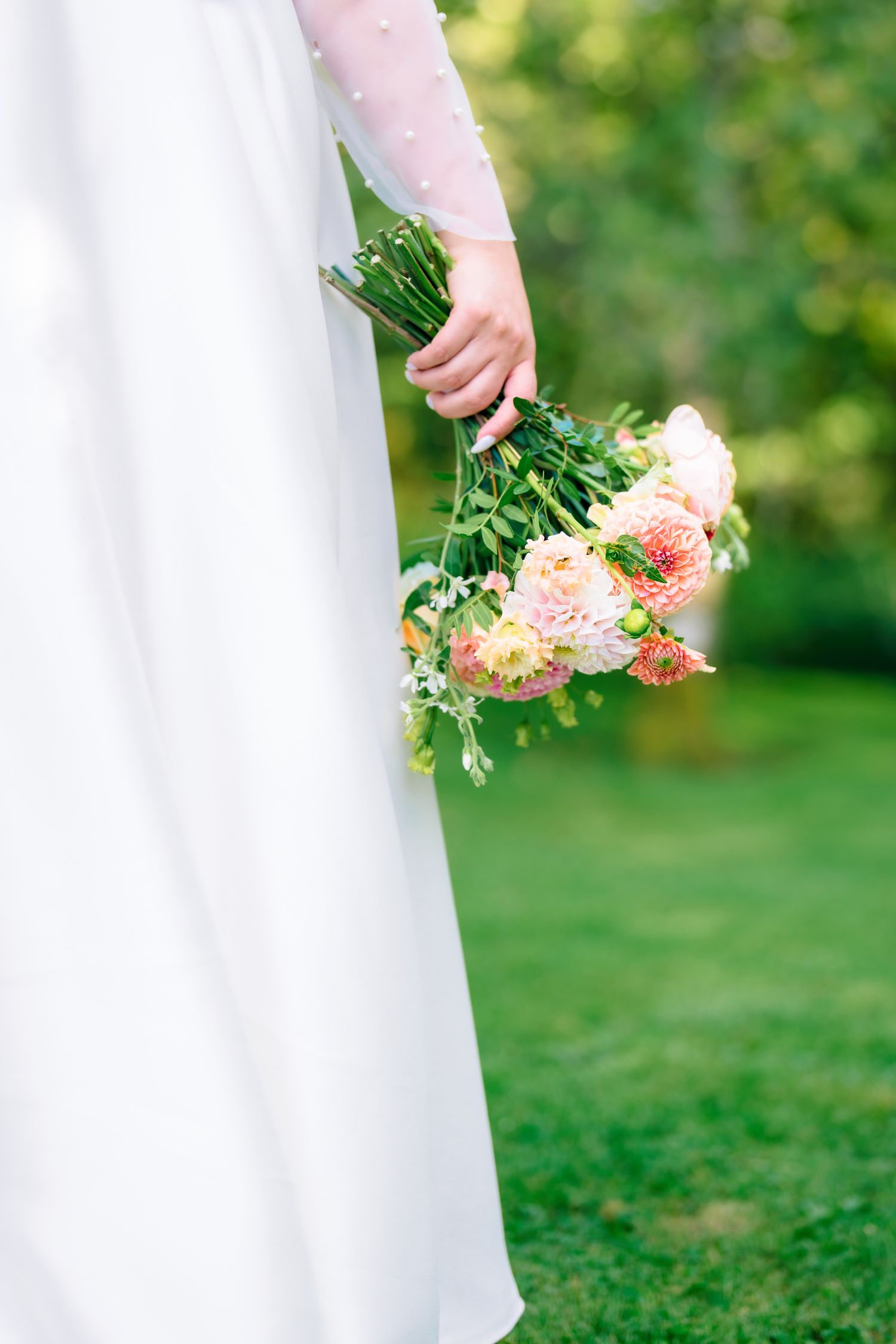 Bride in white dress holding a pastel pink and coral wedding bouquet of dahlias and greenery outdoors.