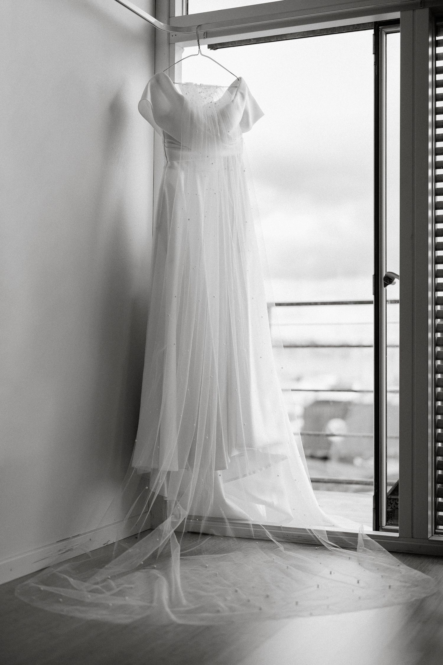 Elegant white wedding dress with flowing veil hanging on a hanger in a window in black and white.