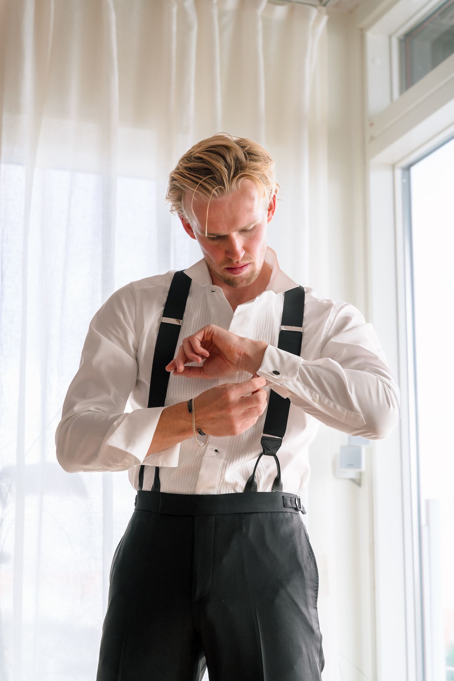 Handsome blond groom in white shirt and suspenders adjusting his cufflinks while getting ready for wedding.