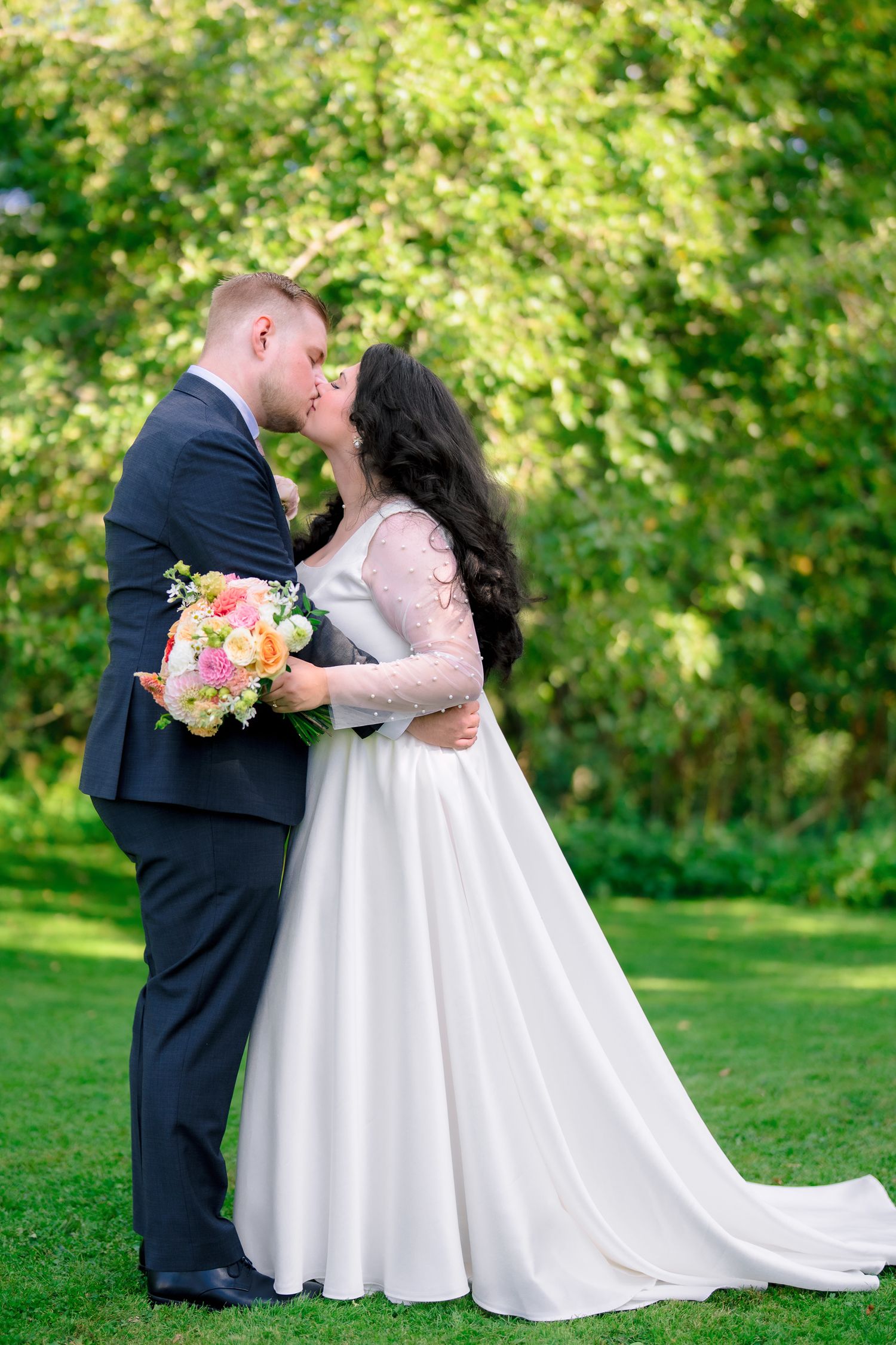 Bride and groom kissing in lush green garden, bride holding colorful bouquet.