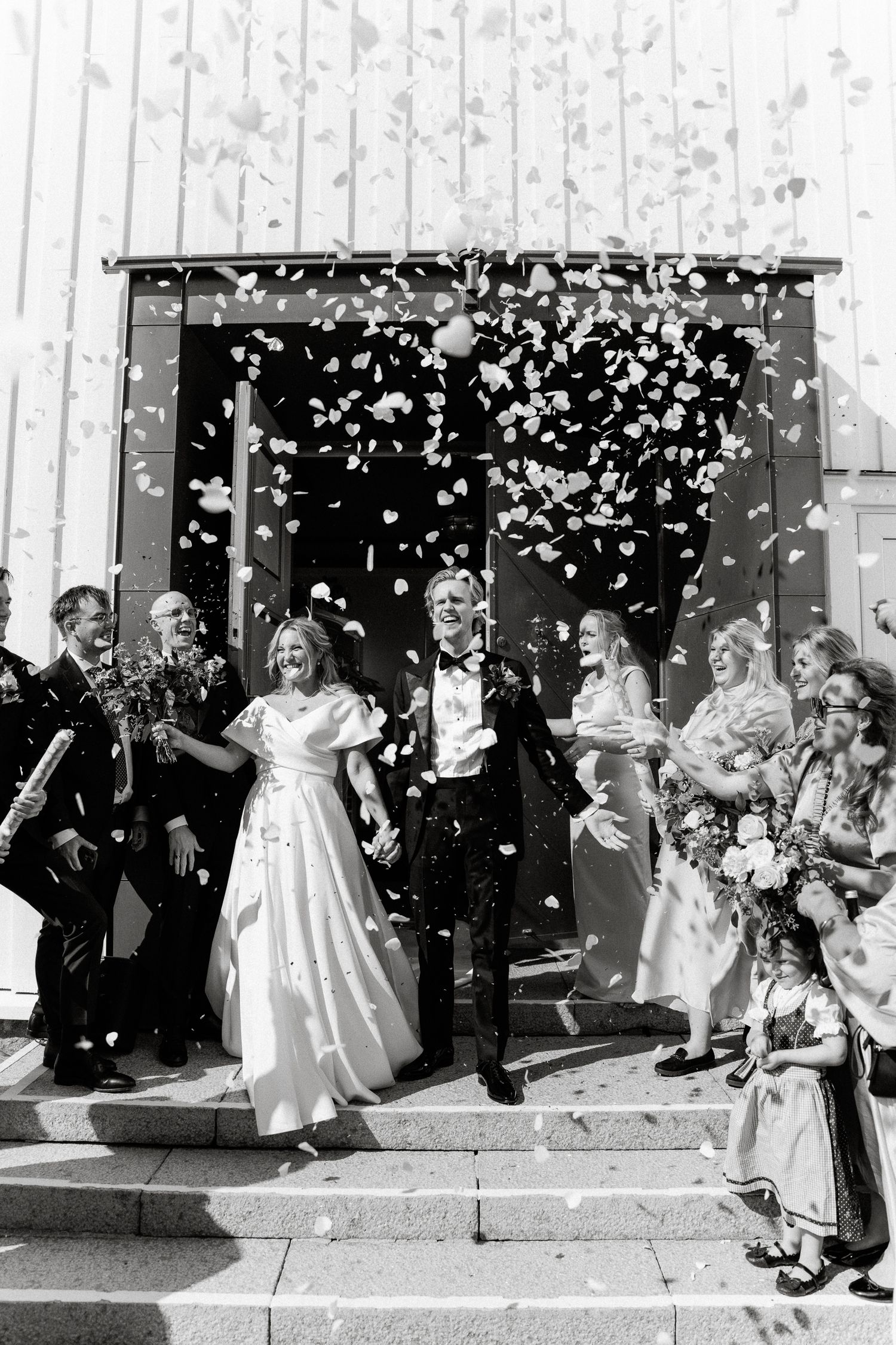 Black and white photo of joyful newlyweds exiting church amid confetti shower with wedding guests celebrating.