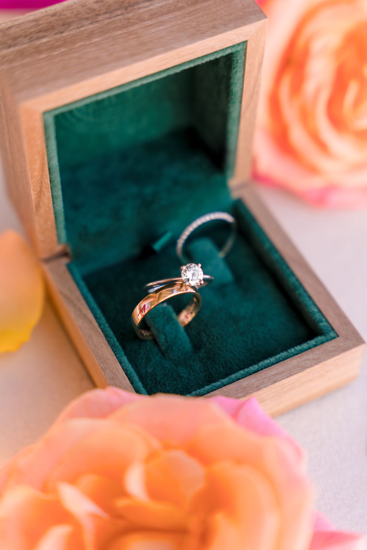 Gold diamond engagement ring and wedding band displayed in a wooden box with green velvet lining, surrounded by peach roses.