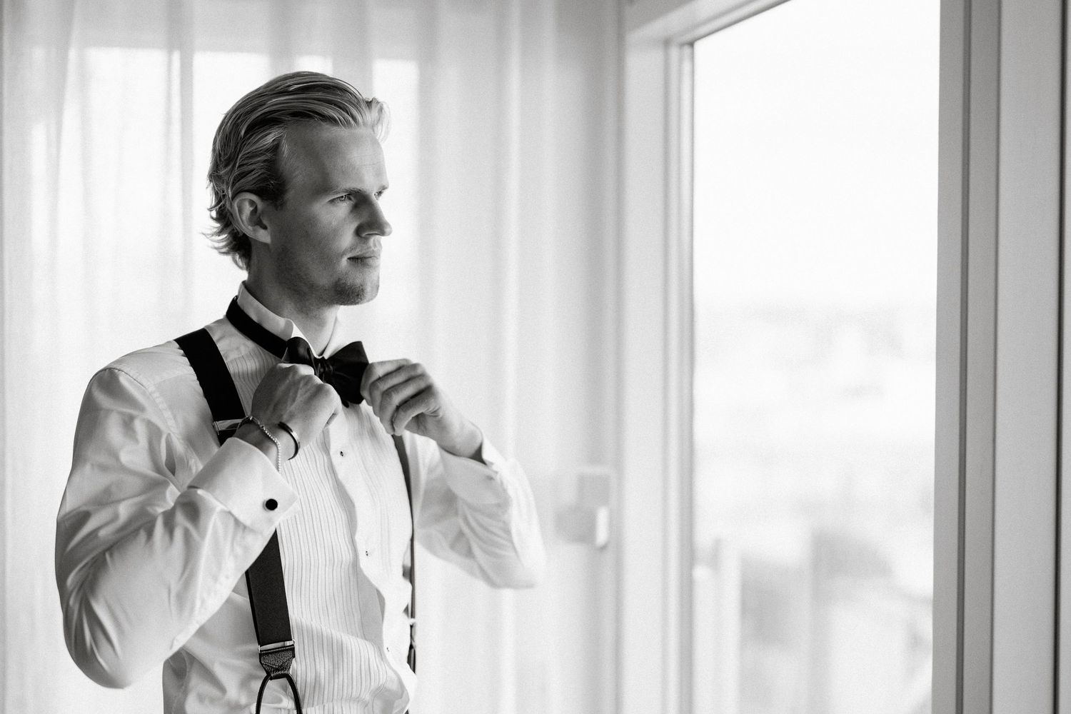 Groom adjusting bow tie wearing suspenders in black and white wedding photo by window.