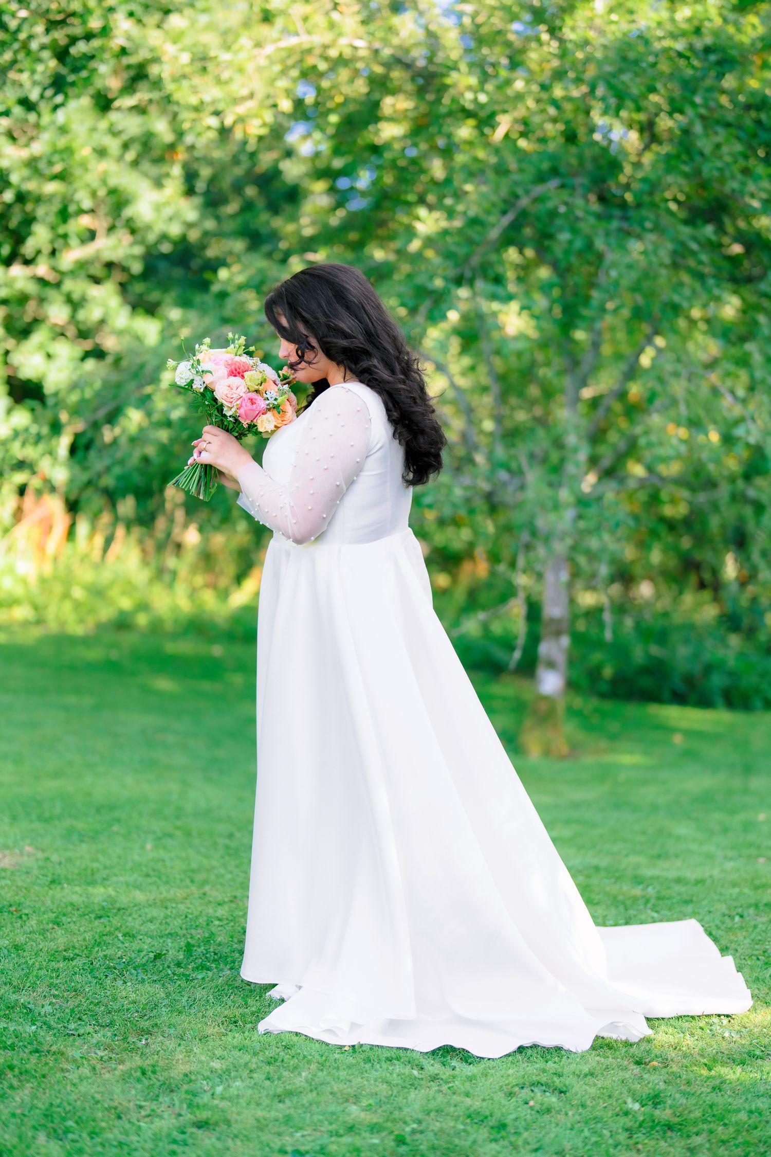 Bride in white long-sleeve gown smelling colorful bouquet in lush green garden.