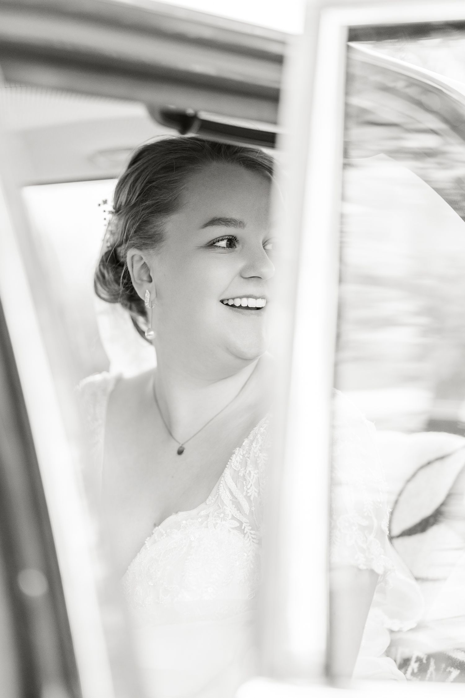 Black and white photo of a smiling bride in a lace dress sitting in a car on her wedding day.
