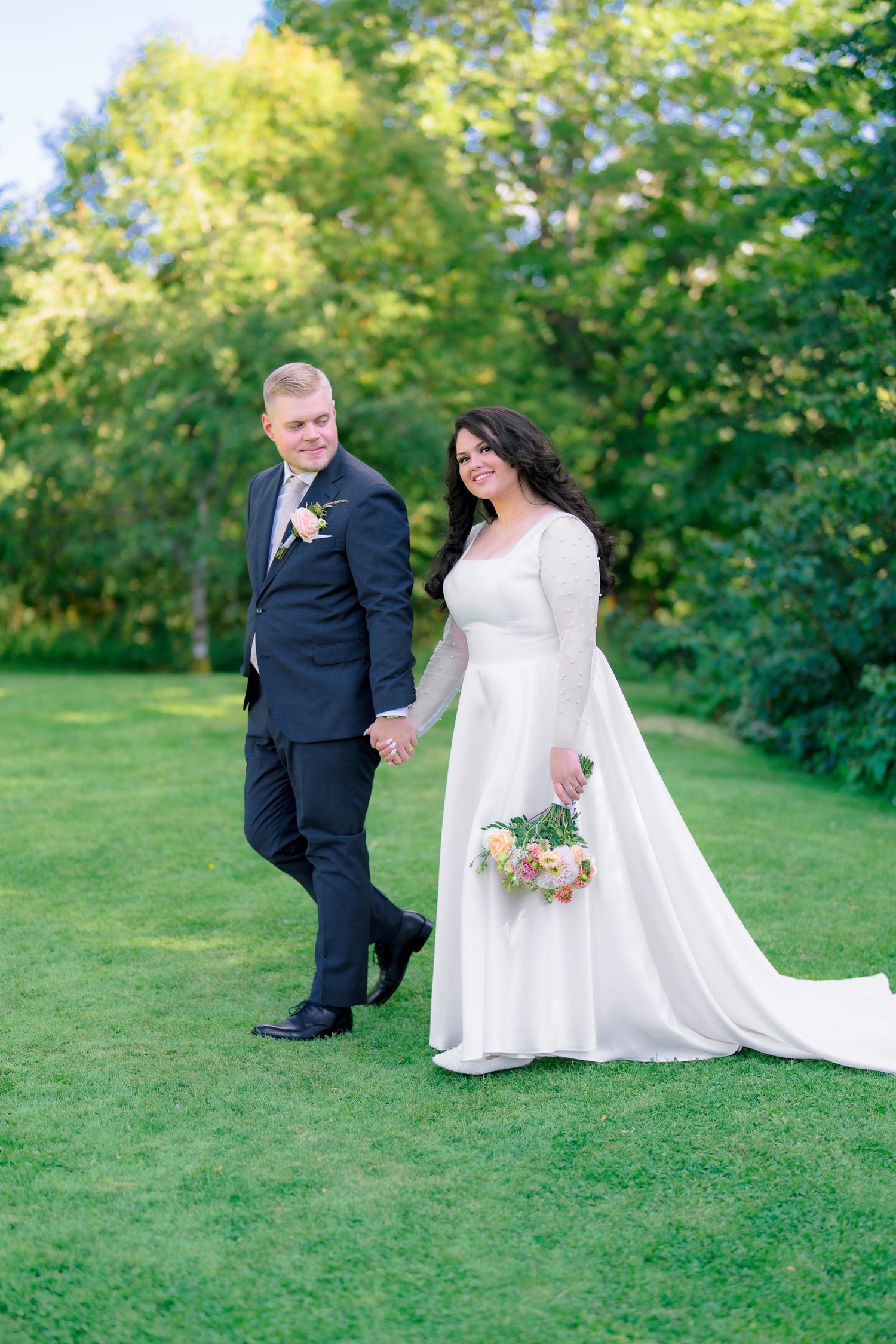 Bride and groom walk hand-in-hand on green lawn, she holds bouquet in white gown, he wears dark suit.