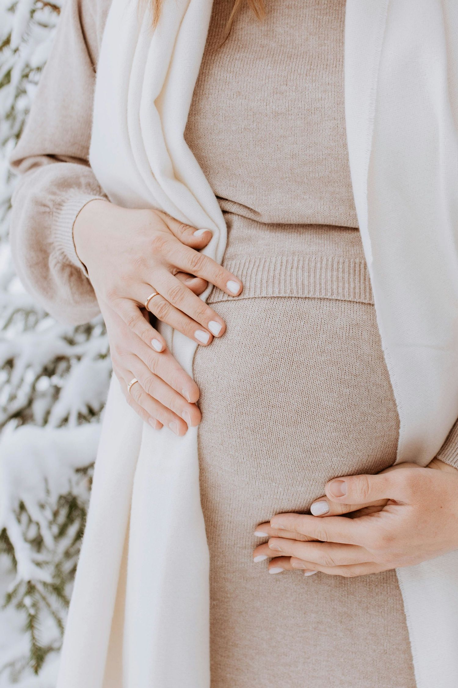 Pregnant woman in beige knit dress holding baby bump outdoors in snowy winter setting.