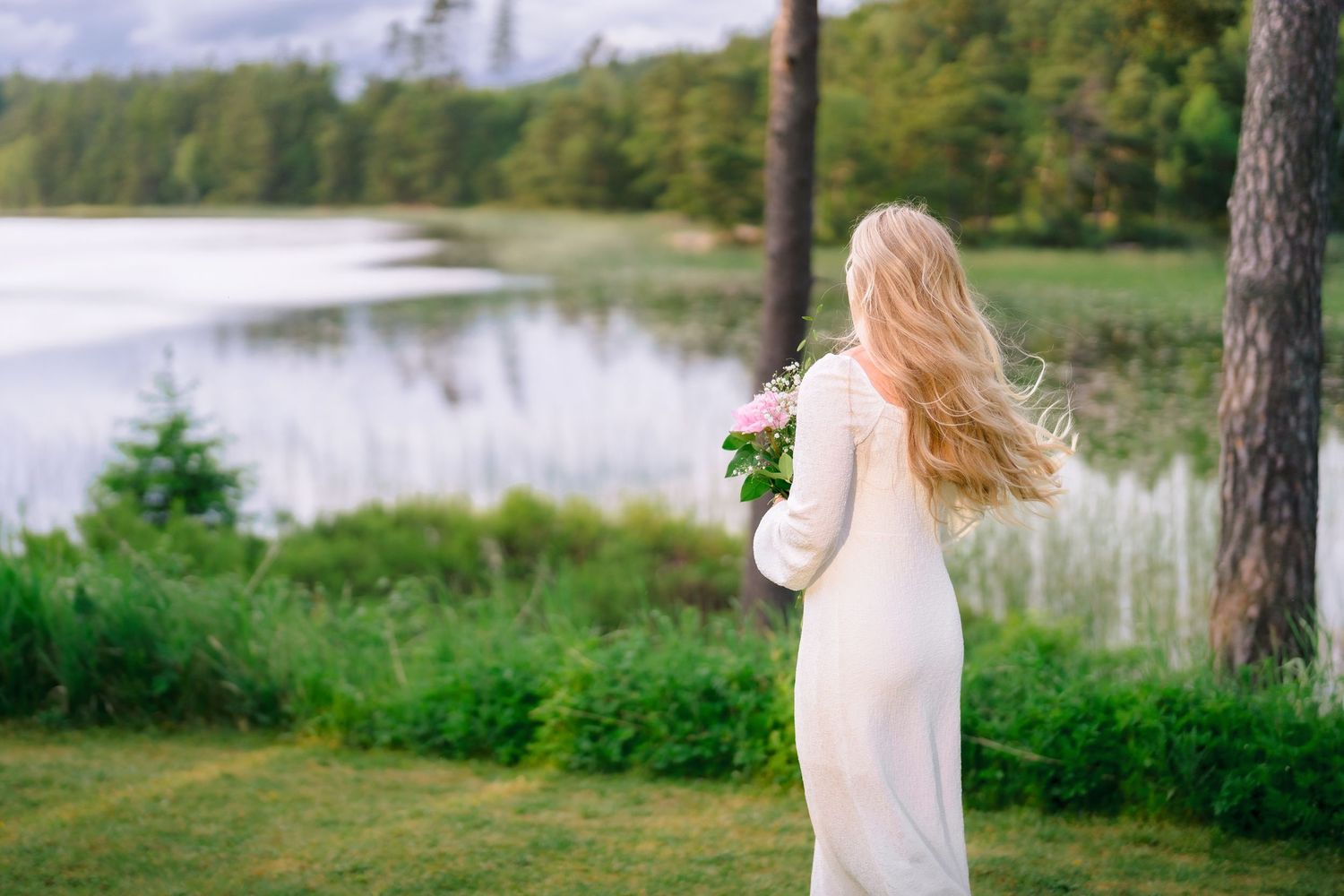 Blonde bride in white gown holding pink bouquet gazing at serene lakeside view.