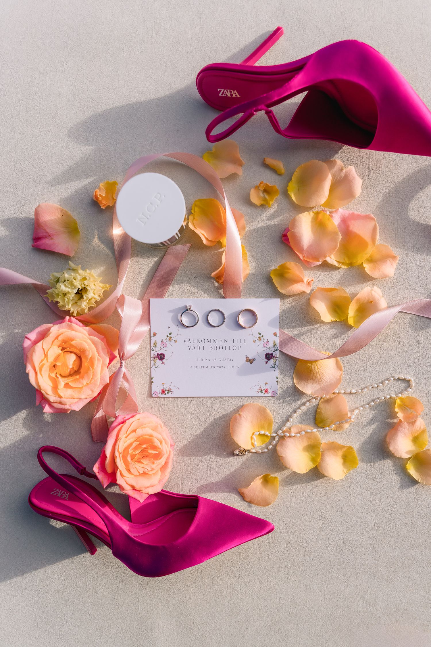 Flat lay of wedding details including pink heels, rings, pearl necklace, roses, and petals on a white surface.