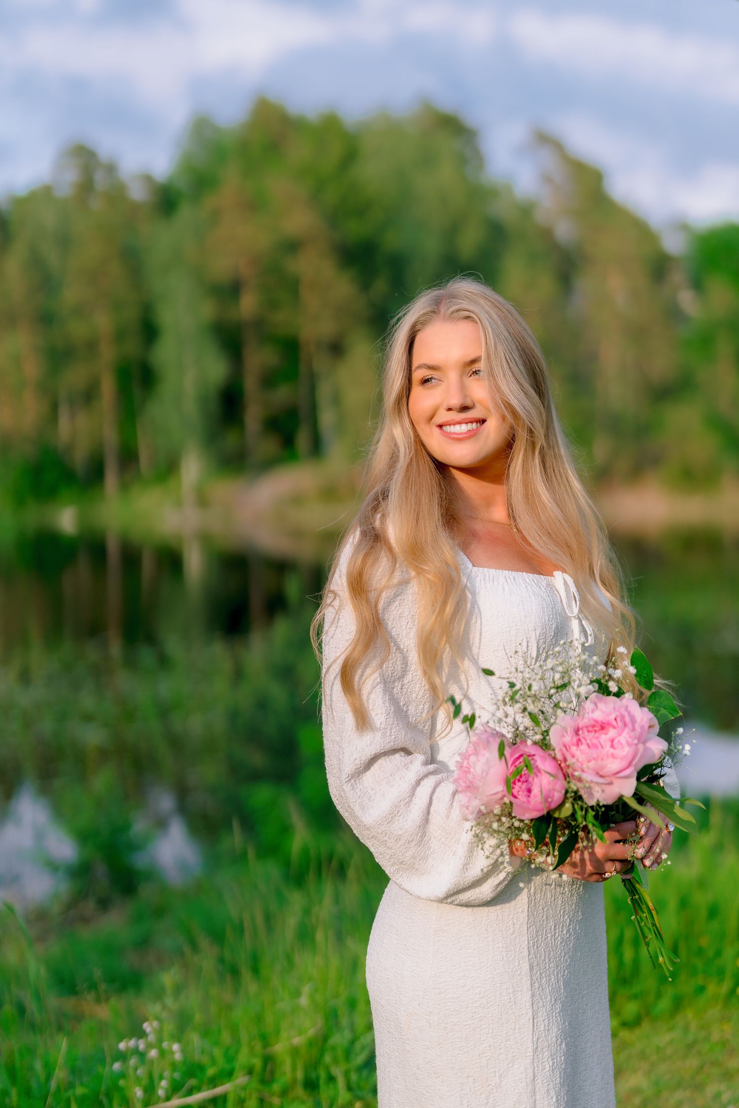 Smiling blonde bride in white dress holding pink peony bouquet with lake backdrop.