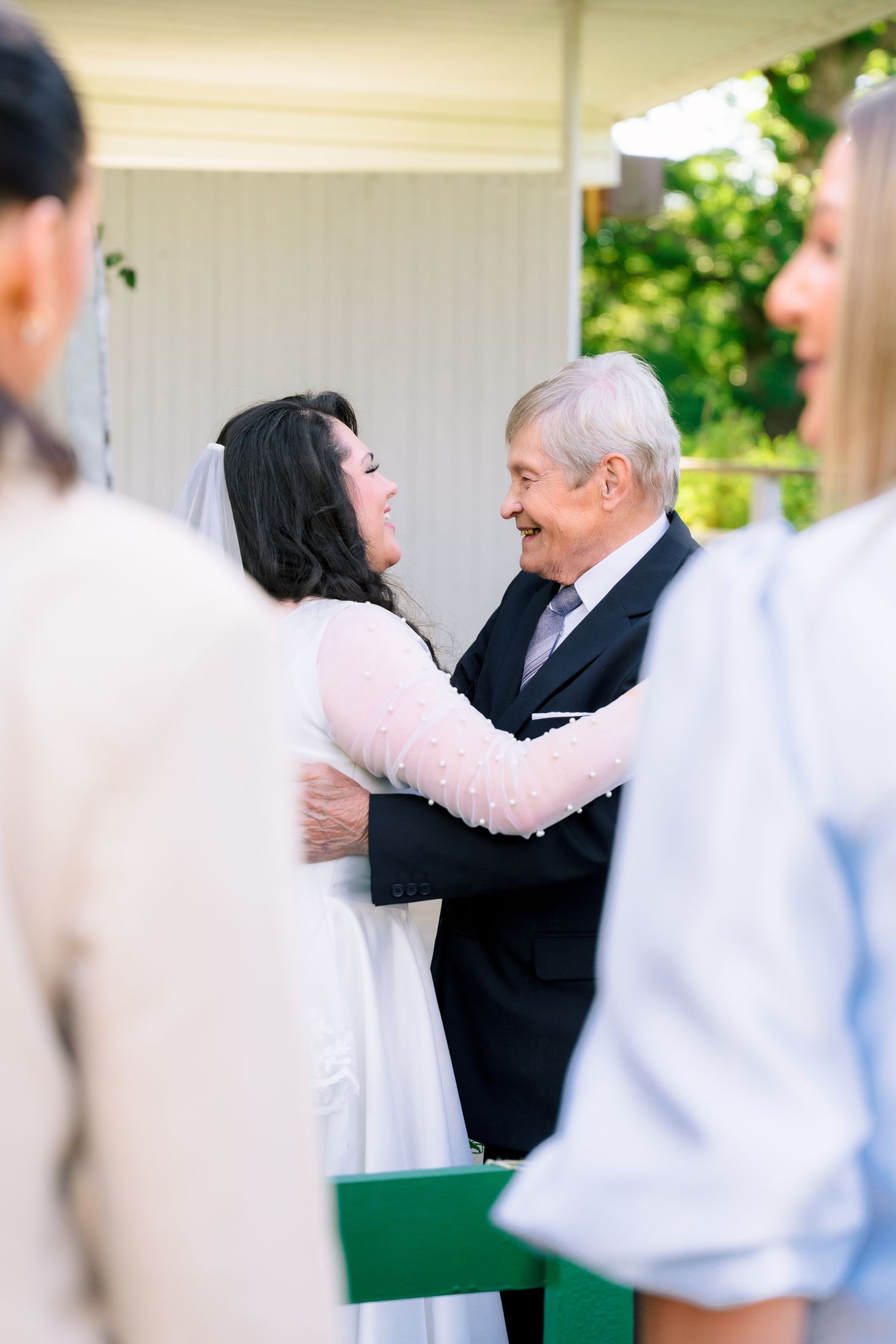 Bride in white gown embracing older man at outdoor wedding ceremony.