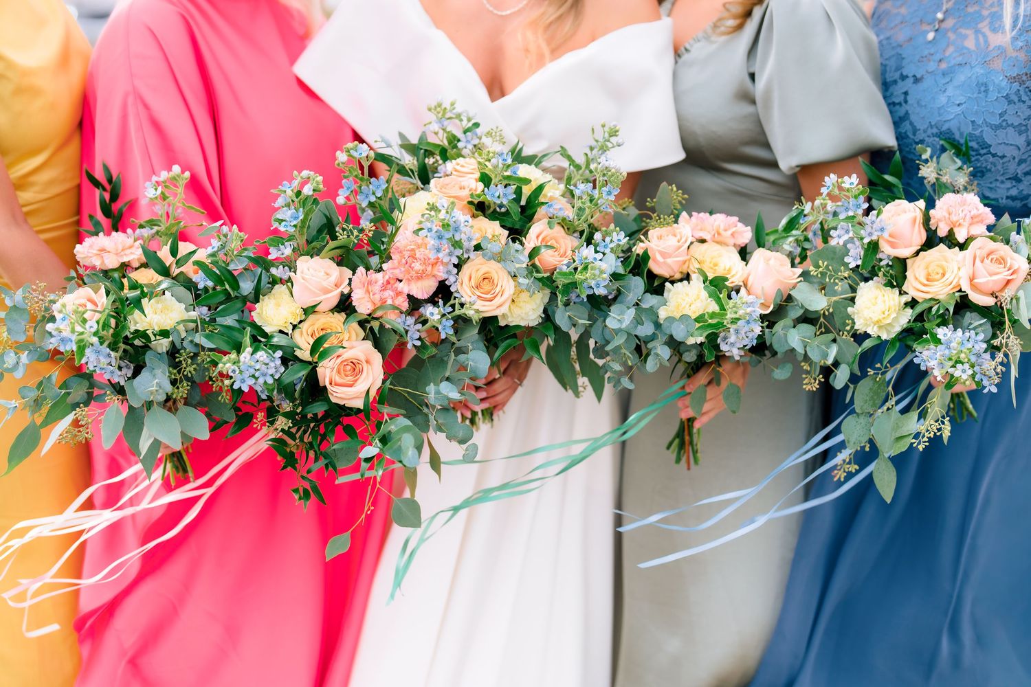 Bridesmaids in colorful dresses holding lush peach rose and greenery bouquets with flowing ribbons.