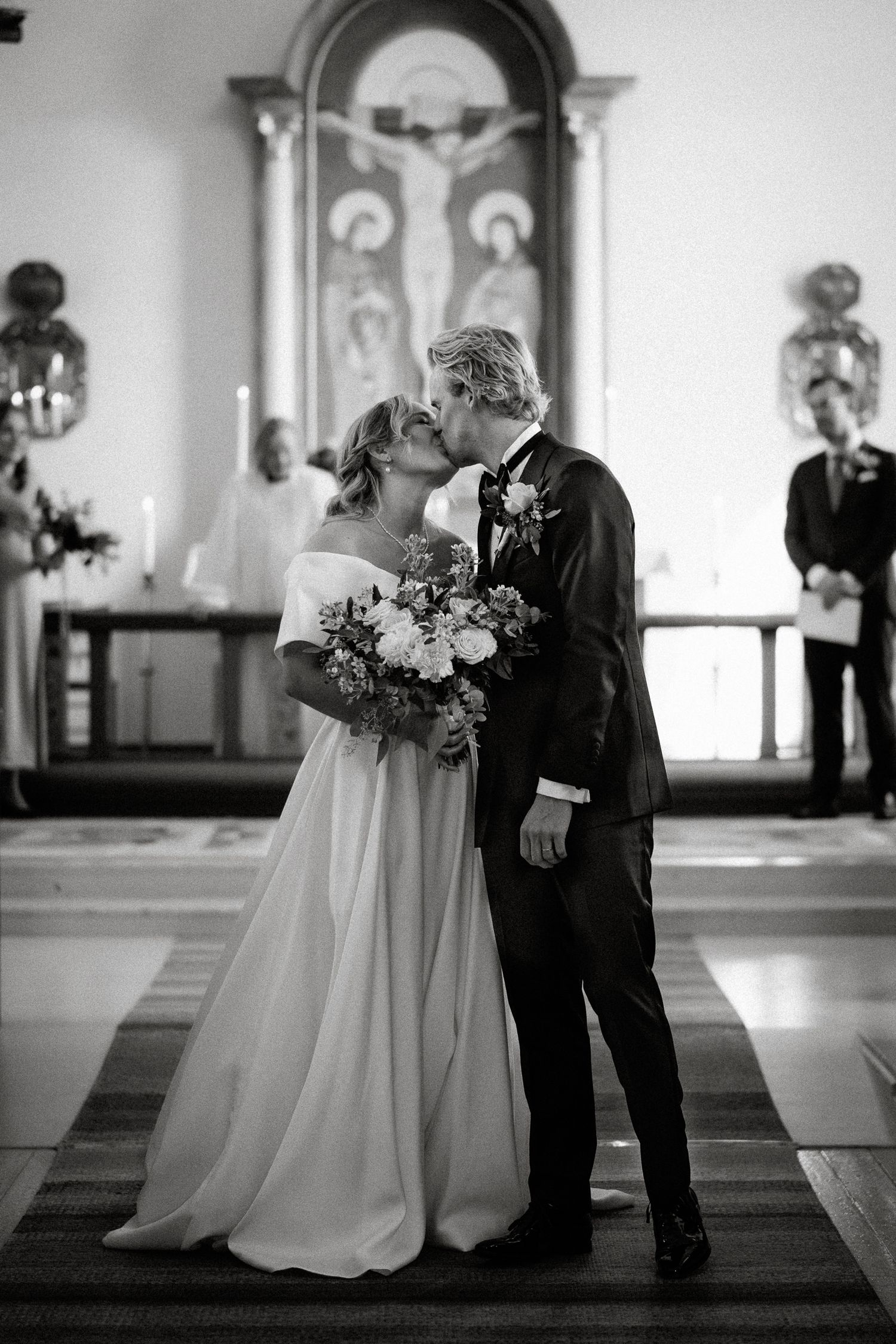 Black and white photo of bride and groom sharing first kiss at church altar during wedding ceremony.