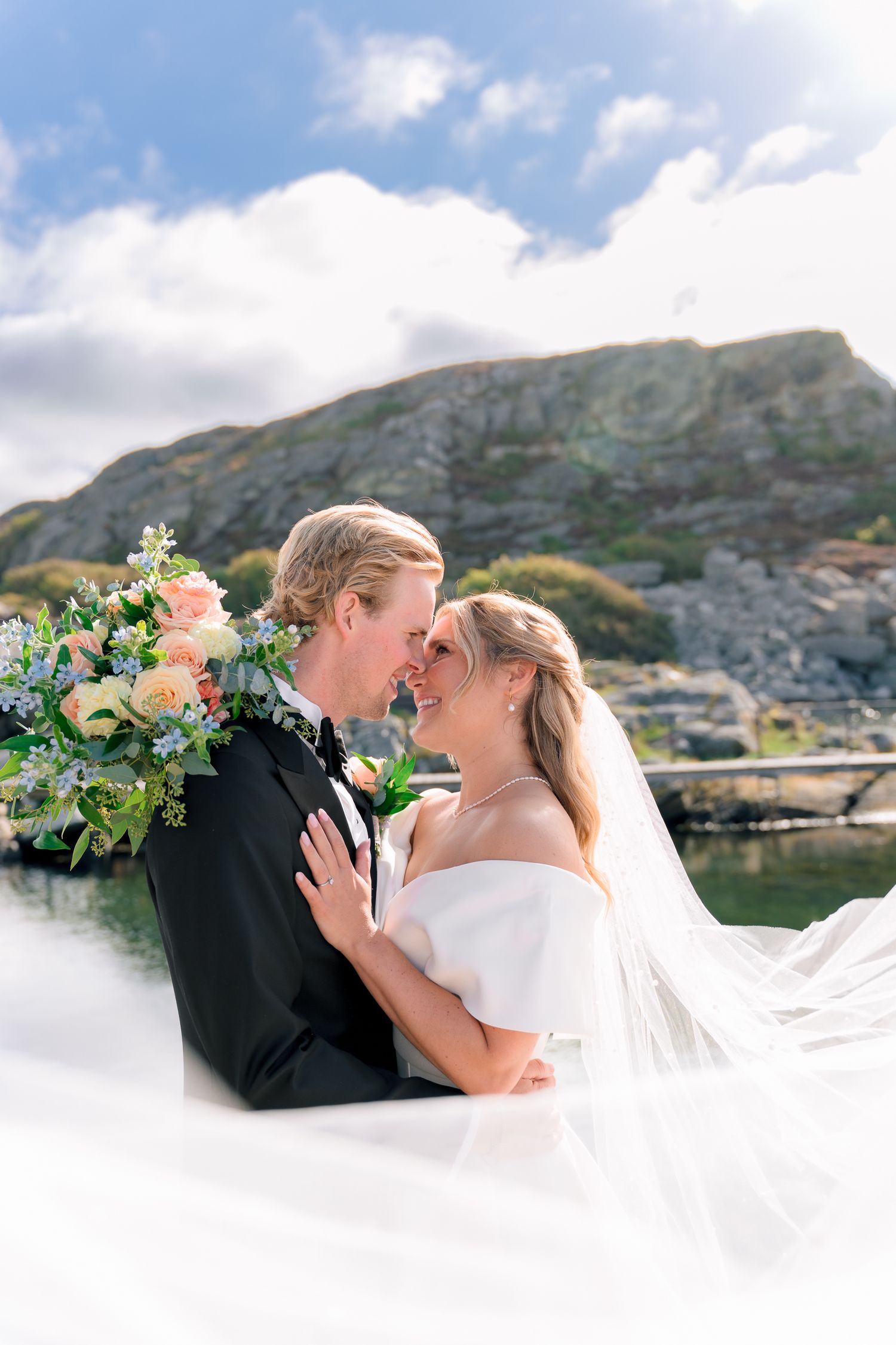 Bride and groom touch noses lovingly as her veil flows beside a scenic waterfront rocky landscape.