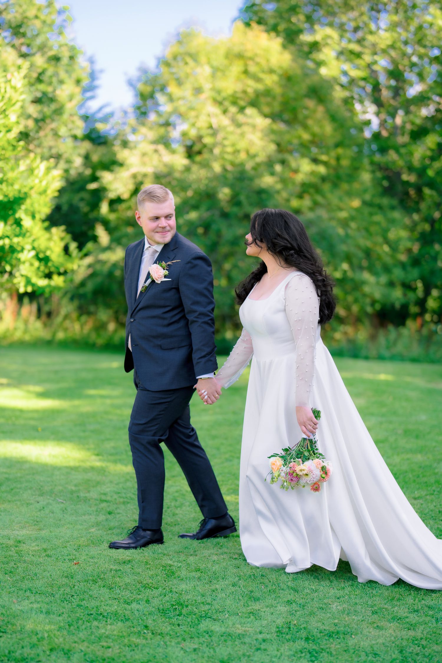 Smiling bride in white gown and groom in black suit holding hands outdoors.