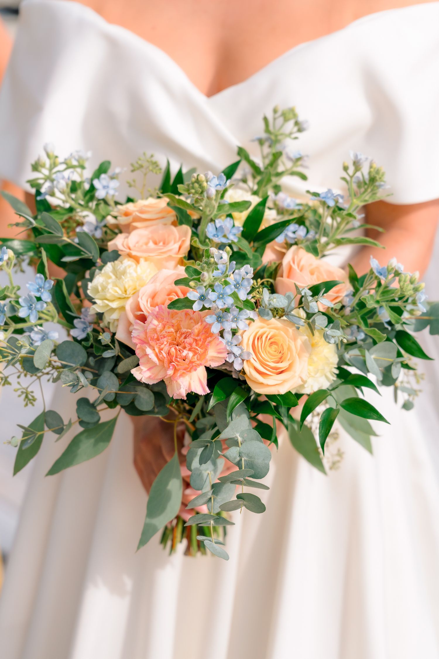 Bride holding a lush wedding bouquet of peach roses, carnations, blue forget-me-nots, and eucalyptus greenery.