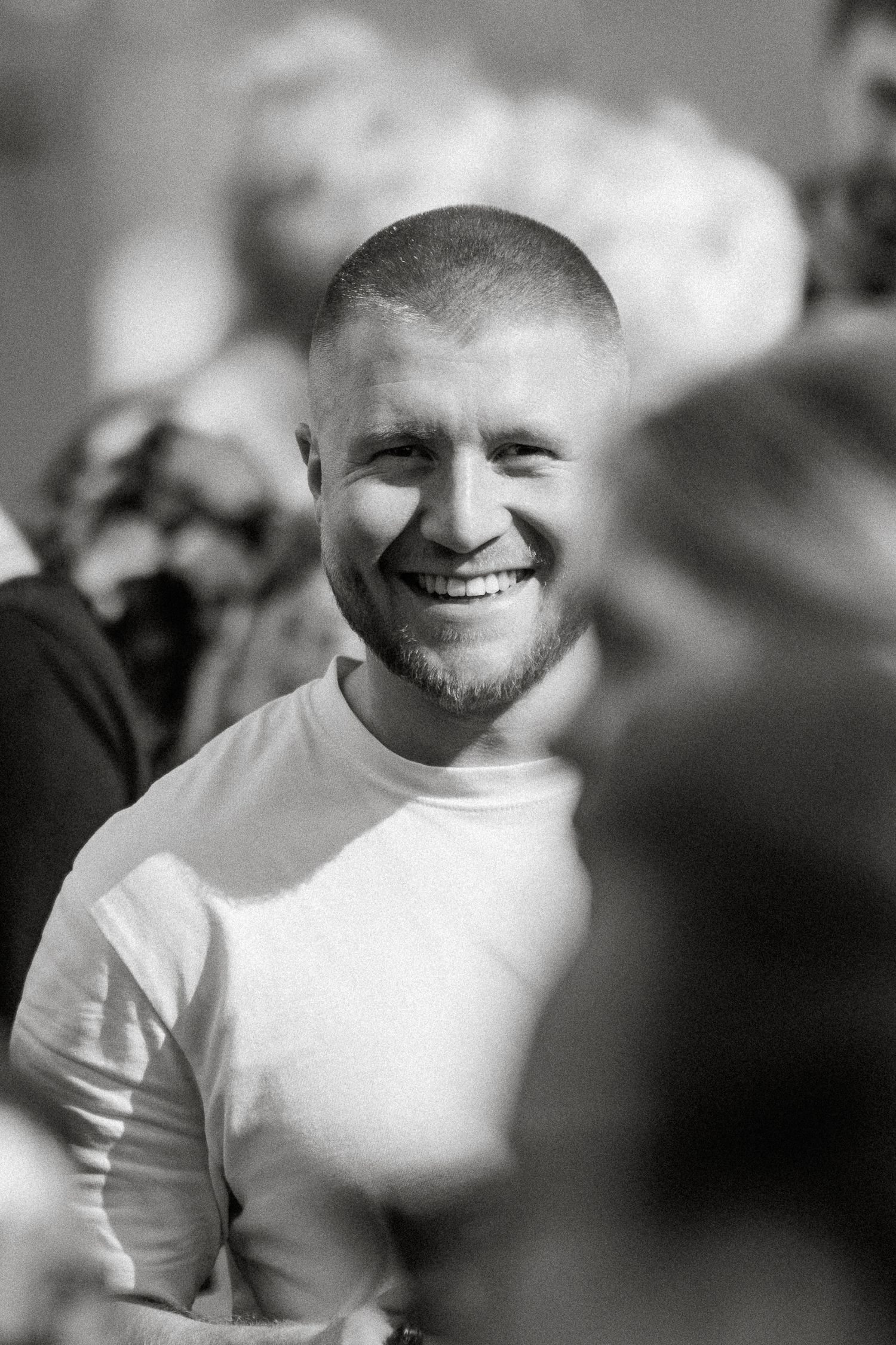 Black and white photo of a smiling young man with a buzz cut wearing a white shirt surrounded by blurred figures.
