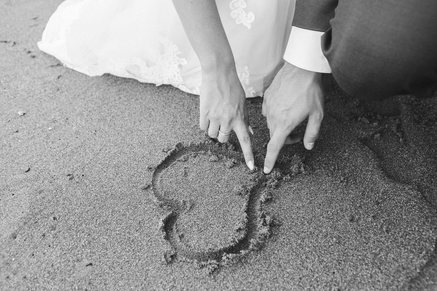 Bride and groom drawing a heart in the sand on a beach in black and white wedding photo.