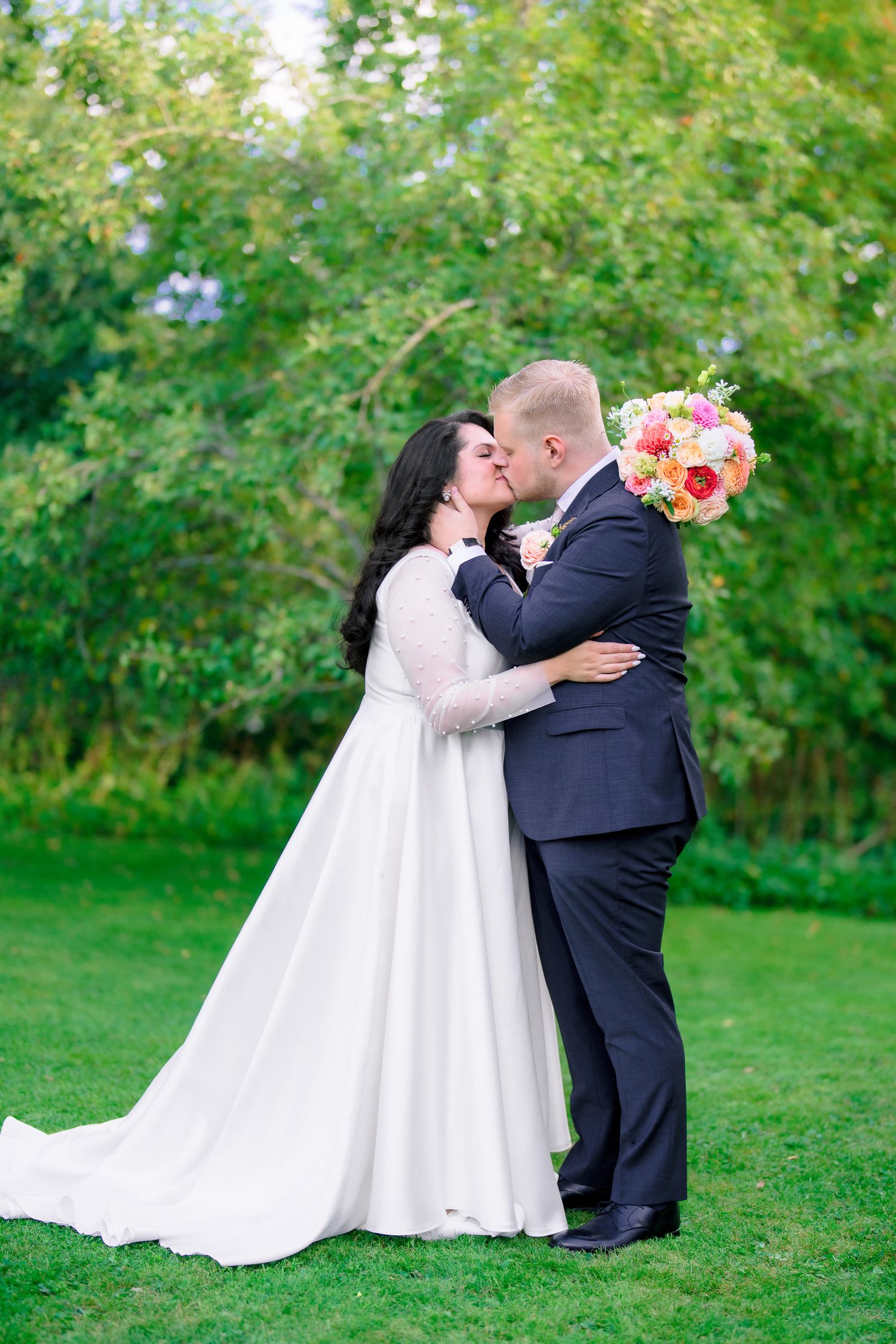 Bride and groom sharing a kiss outdoors holding colorful pink and orange bouquet.