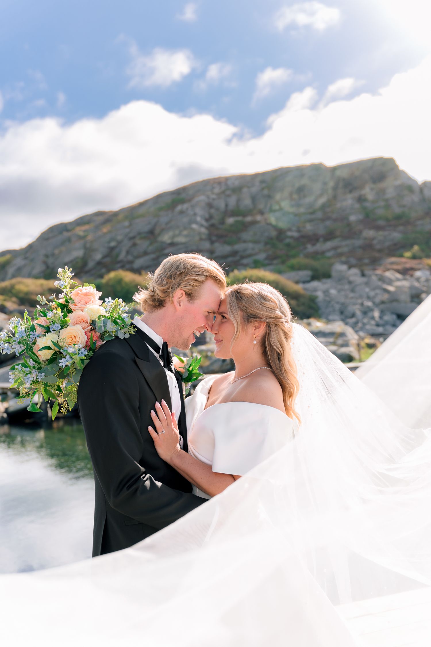 Bride and groom share an intimate forehead touch as her veil sweeps across a rocky waterside setting.