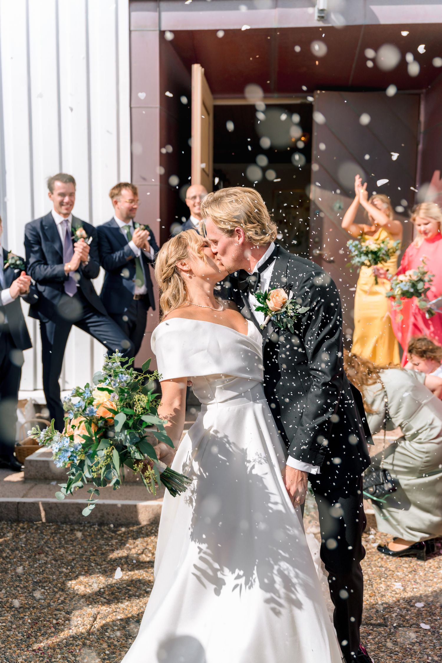 Bride and groom sharing a kiss outside church entrance as guests celebrate with confetti in sunlight.