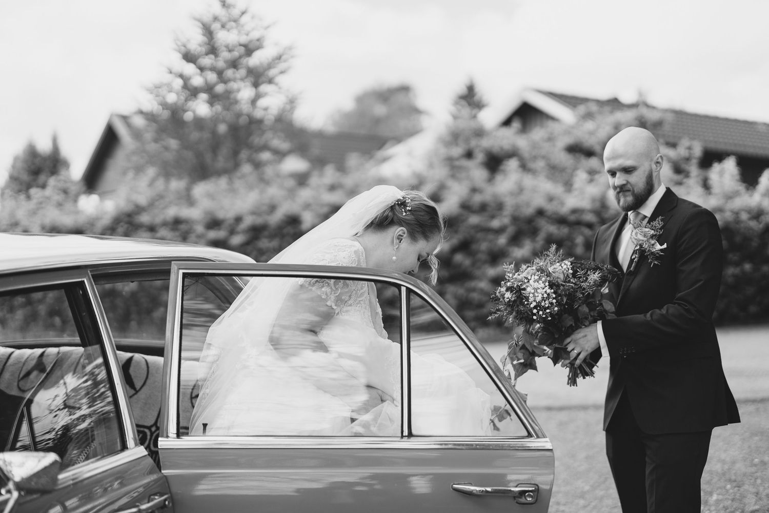 Groom helping bride exit vintage car on wedding day in black and white.
