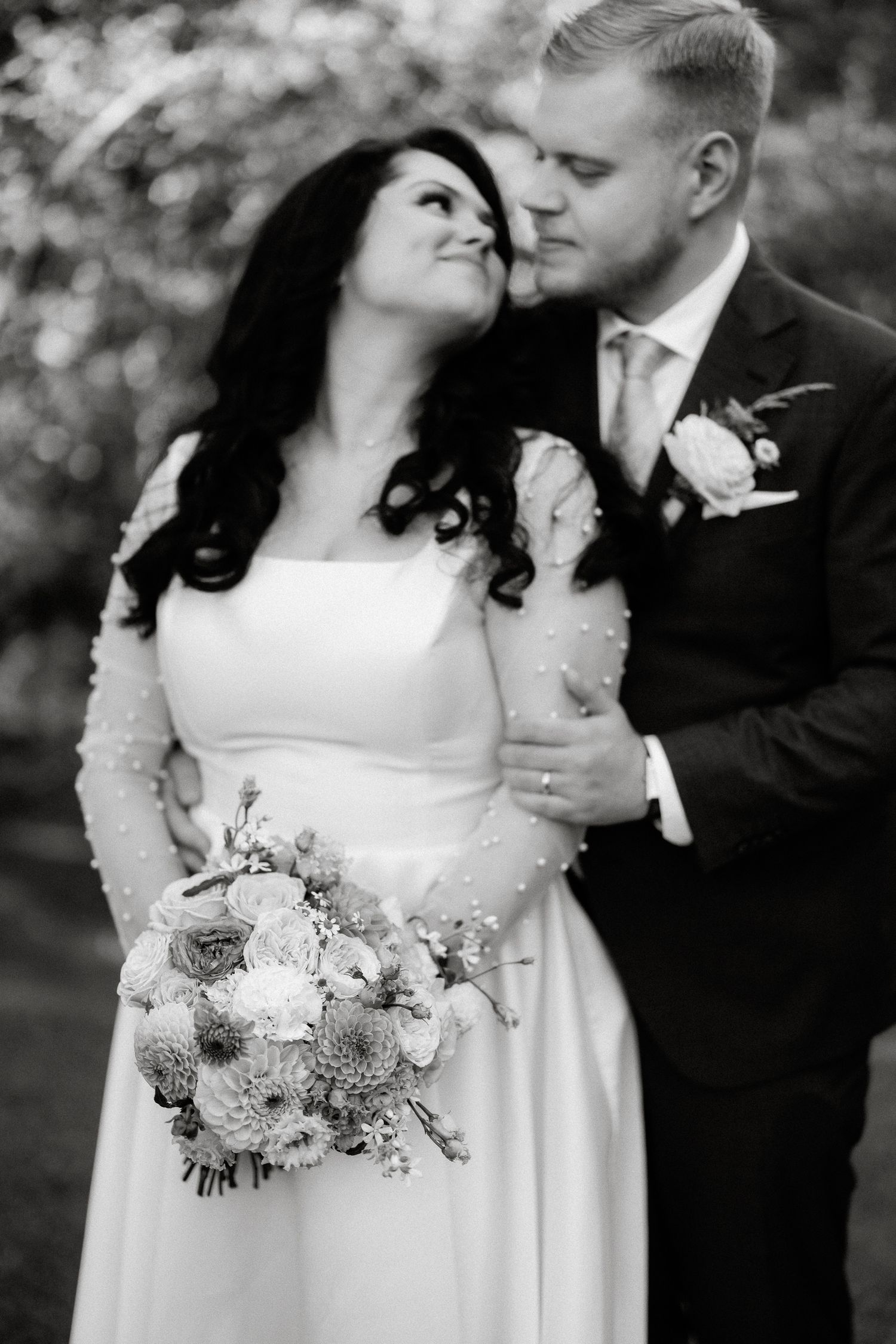Black and white portrait of bride and groom embracing with floral bouquet outdoors.